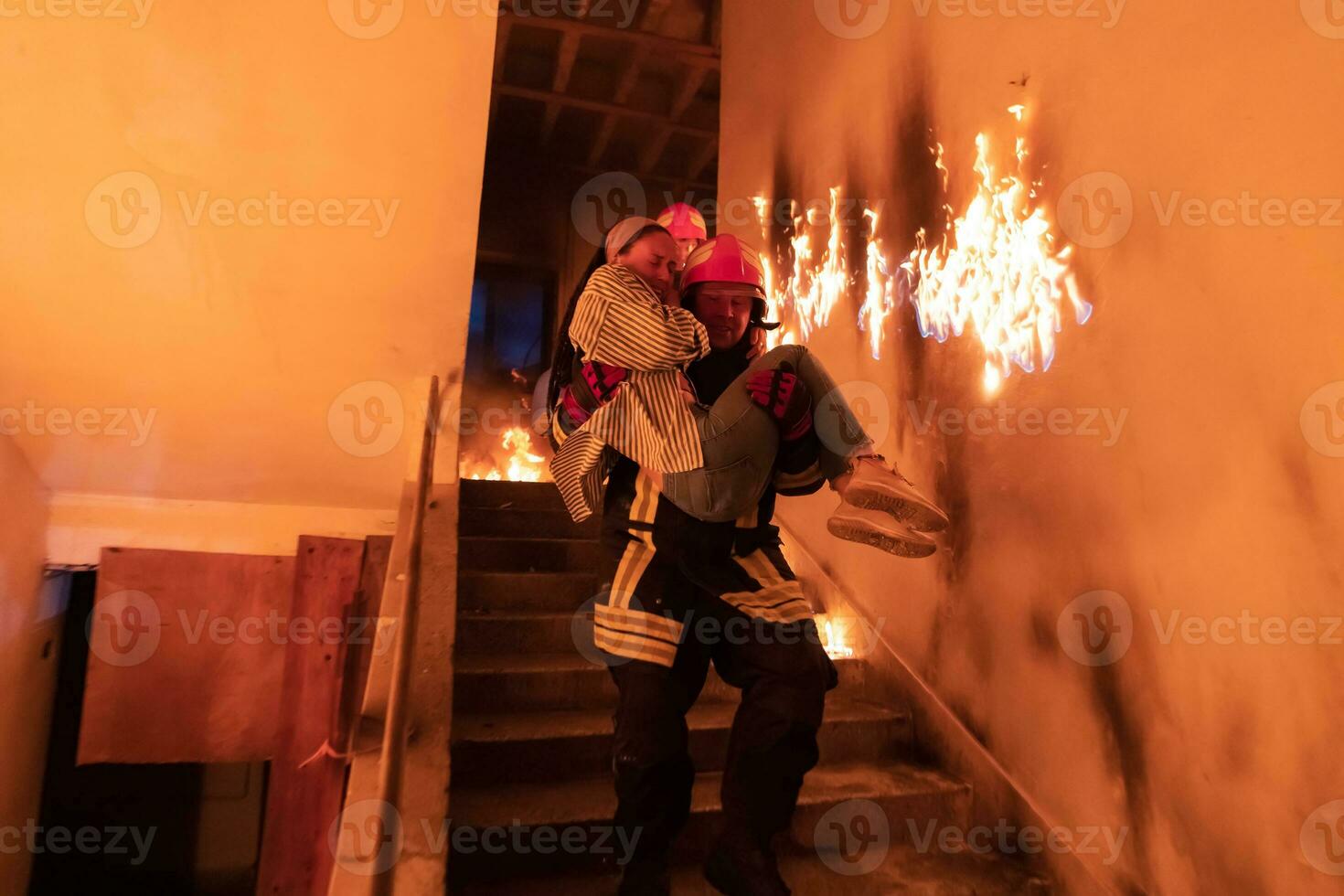 Brave Fireman Descends Stairs of a Burning Building and Holds Saved Girl in His Arms. Open fire and one Firefighter in the Background. photo