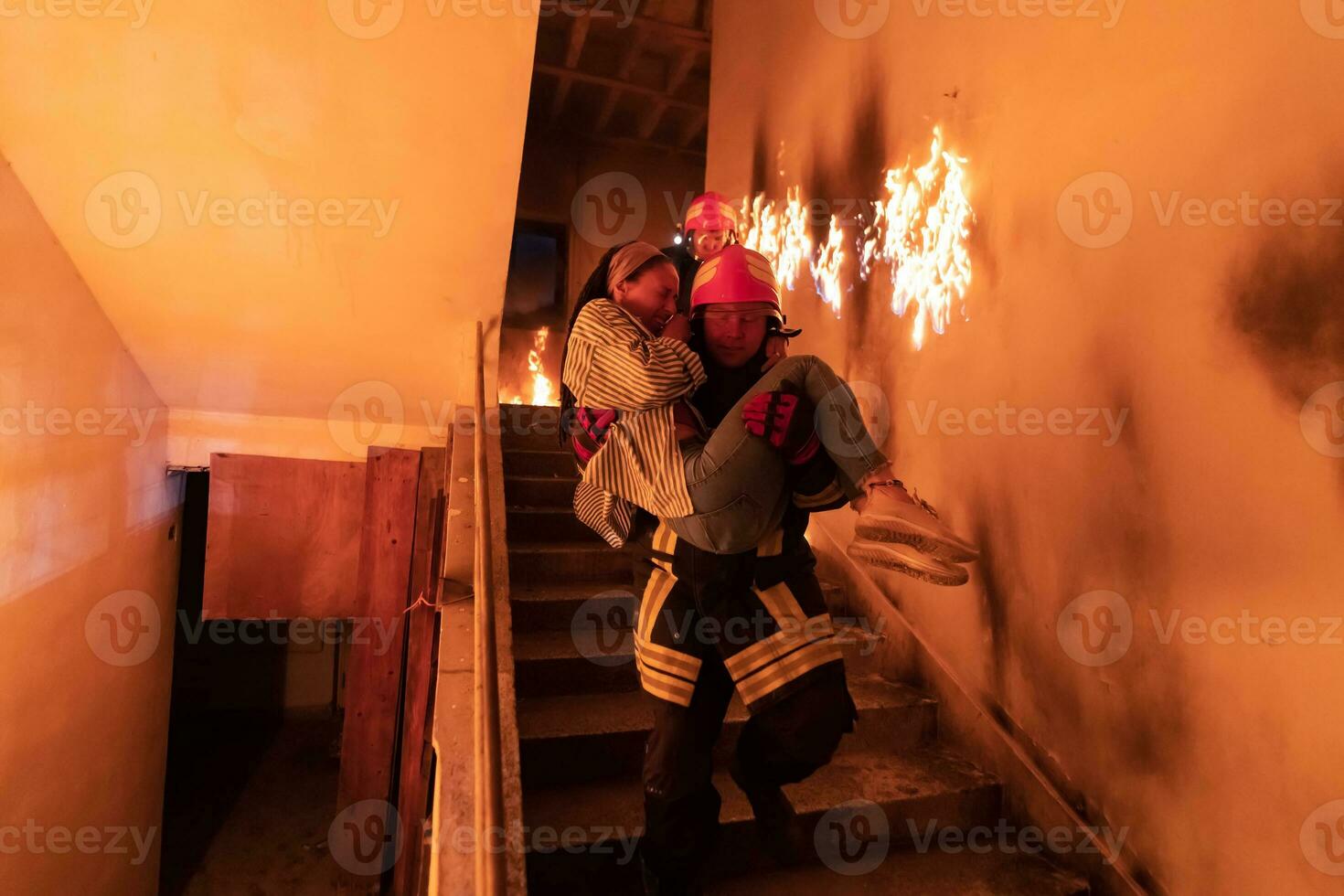 Brave Fireman Descends Stairs of a Burning Building and Holds Saved Girl in His Arms. Open fire and one Firefighter in the Background. photo