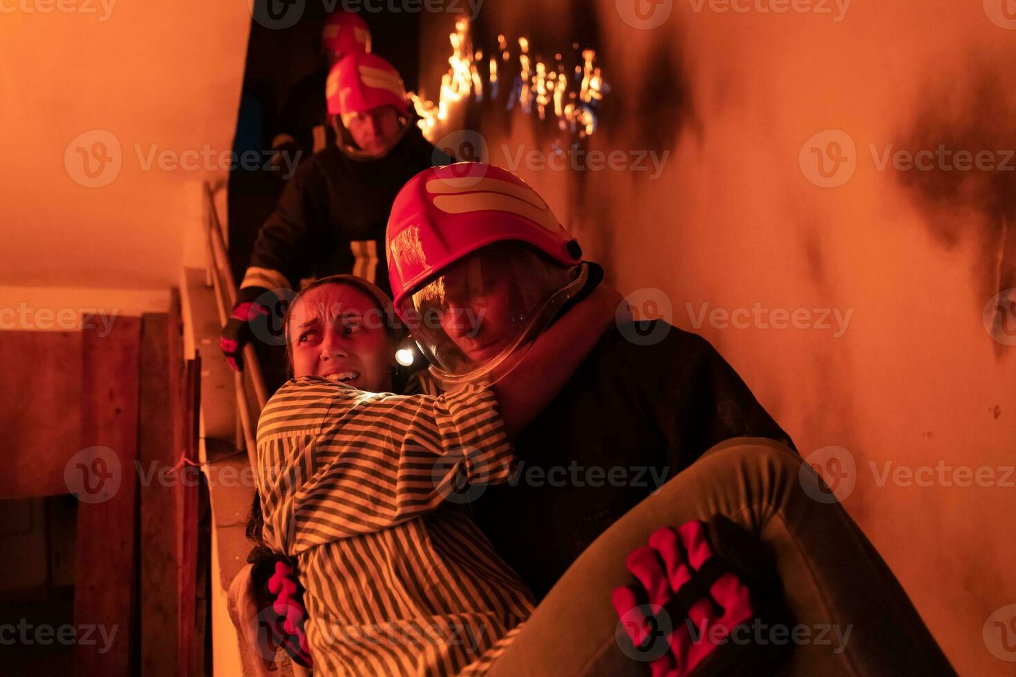 Brave Fireman Descends Stairs of a Burning Building and Holds Saved Girl in His Arms. Open fire and one Firefighter in the Background. photo