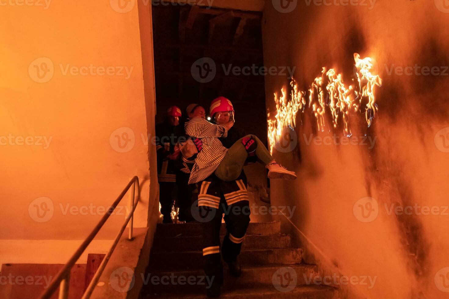 Brave Fireman Descends Stairs of a Burning Building and Holds Saved Girl in His Arms. Open fire and one Firefighter in the Background. photo