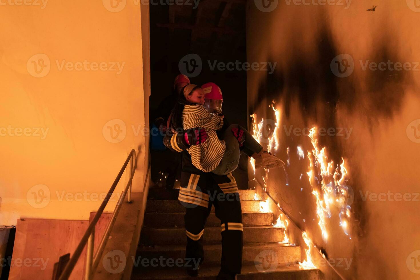 Brave Fireman Descends Stairs of a Burning Building and Holds Saved Girl in His Arms. Open fire and one Firefighter in the Background. photo