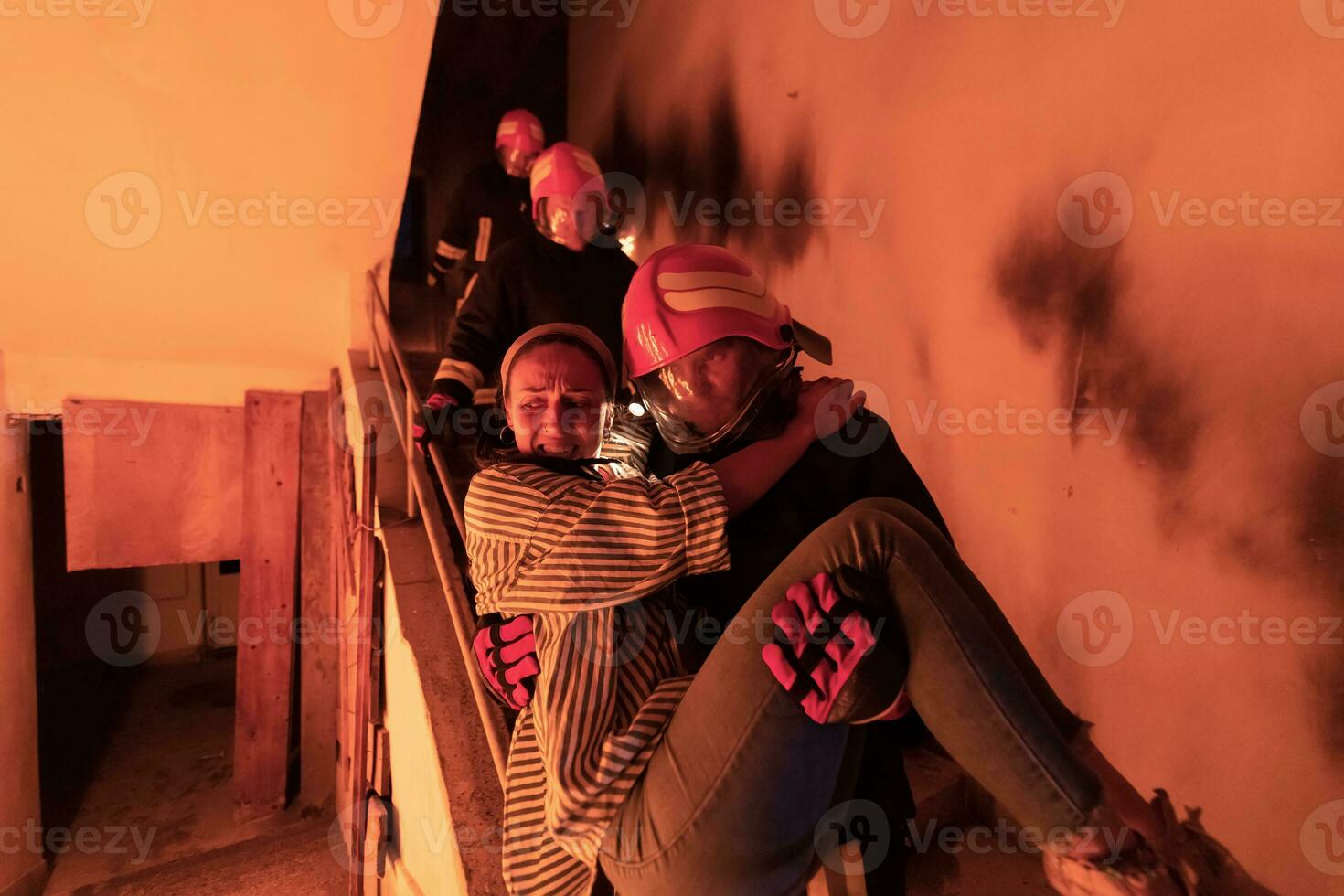 Brave Fireman Descends Stairs of a Burning Building and Holds Saved Girl in His Arms. Open fire and one Firefighter in the Background. photo