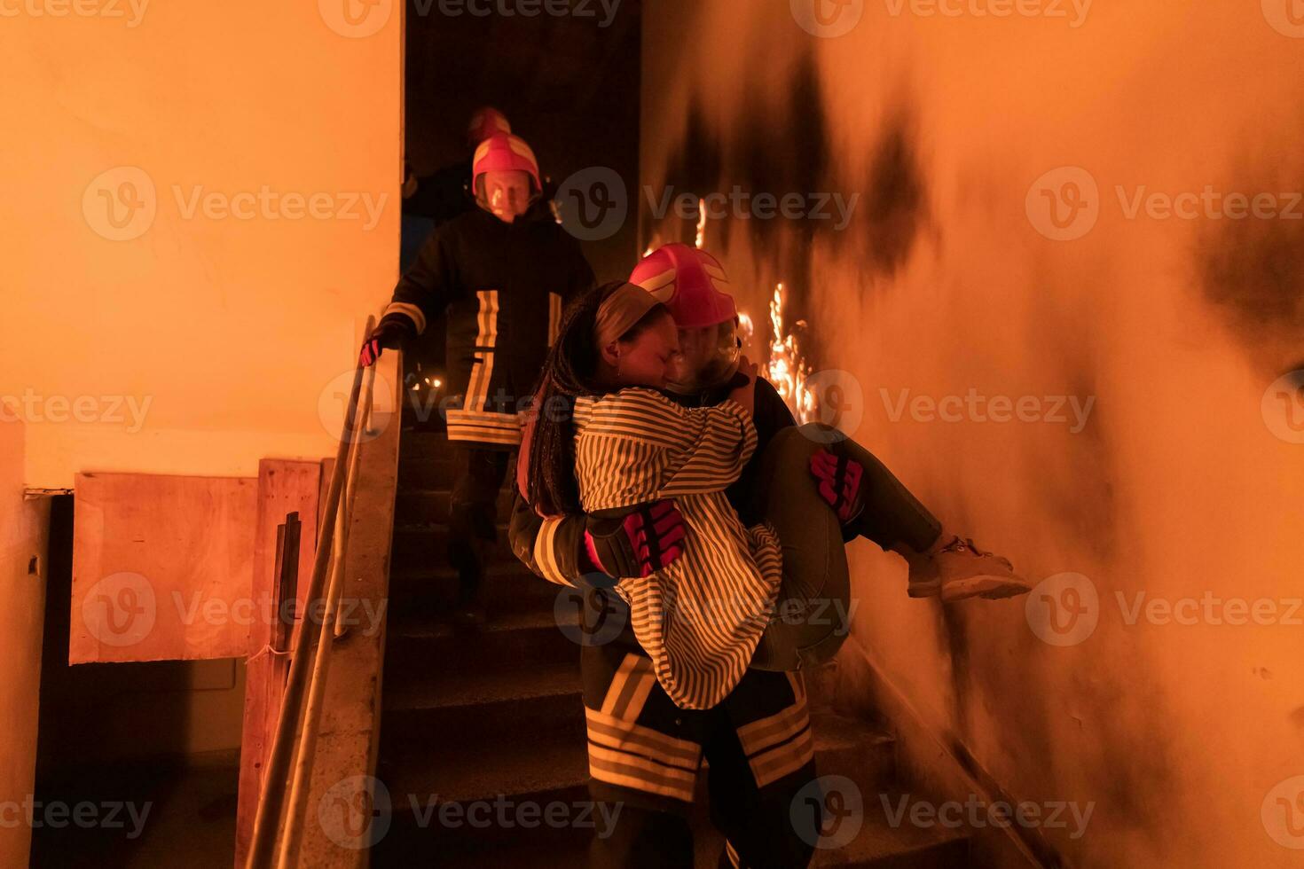 Brave Fireman Descends Stairs of a Burning Building and Holds Saved Girl in His Arms. Open fire and one Firefighter in the Background. photo