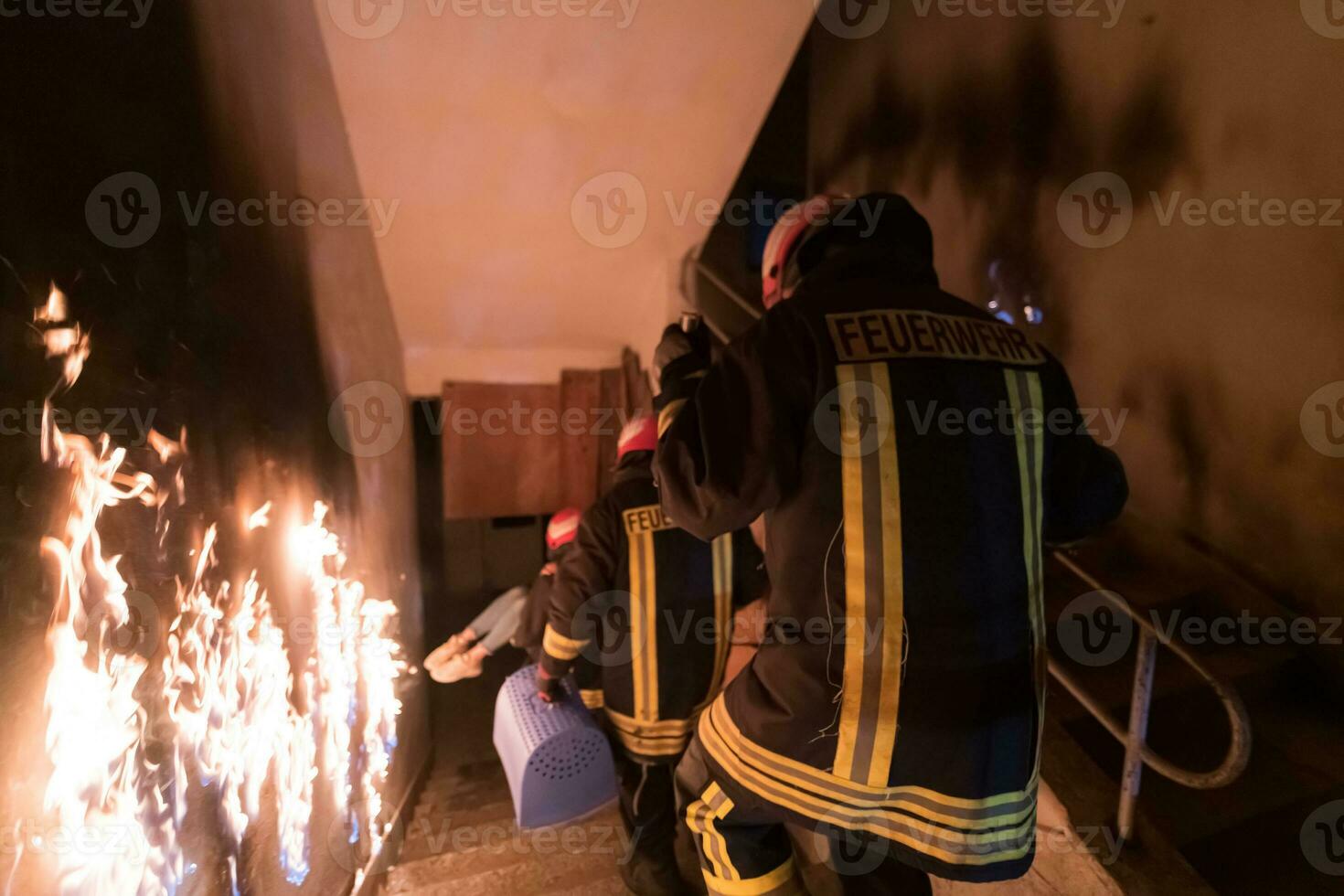 Brave Fireman Descends Stairs of a Burning Building and Holds Saved Girl in His Arms. Open fire and one Firefighter in the Background. photo
