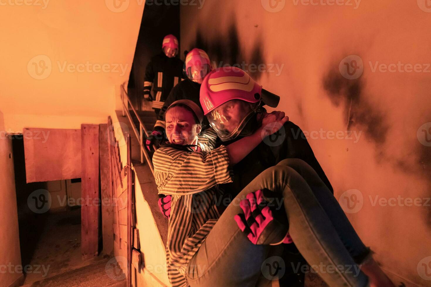 Brave Fireman Descends Stairs of a Burning Building and Holds Saved Girl in His Arms. Open fire and one Firefighter in the Background. photo