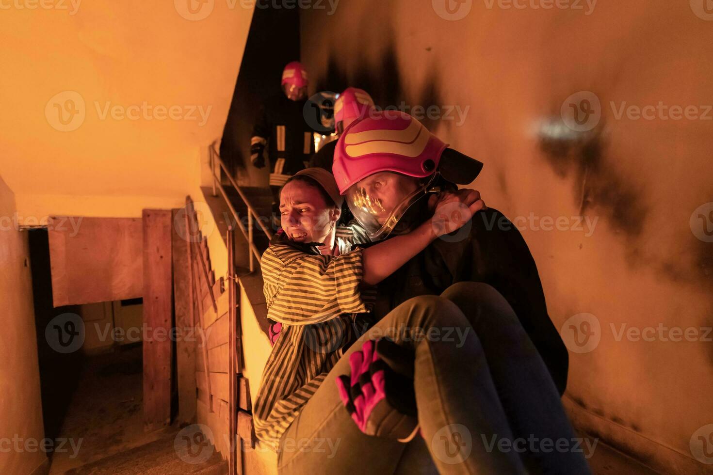 Brave Fireman Descends Stairs of a Burning Building and Holds Saved Girl in His Arms. Open fire and one Firefighter in the Background. photo