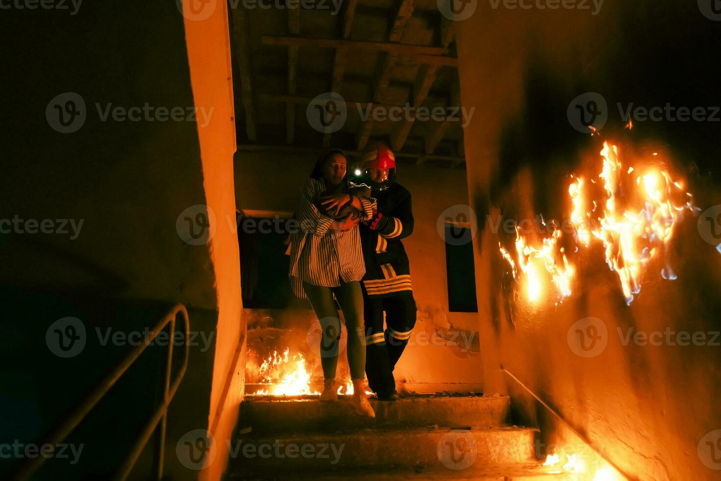 Brave Fireman Descends Stairs of a Burning Building and Holds Saved Girl in His Arms. Open fire and one Firefighter in the Background. photo