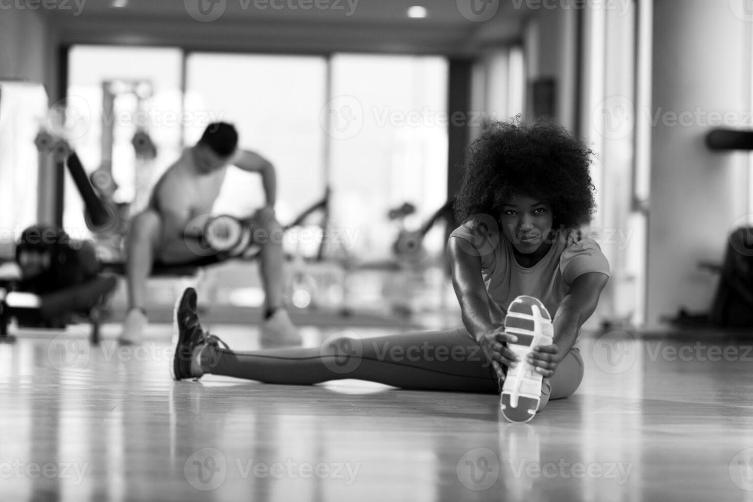 woman in a gym stretching and warming up man in background working with dumbbels photo