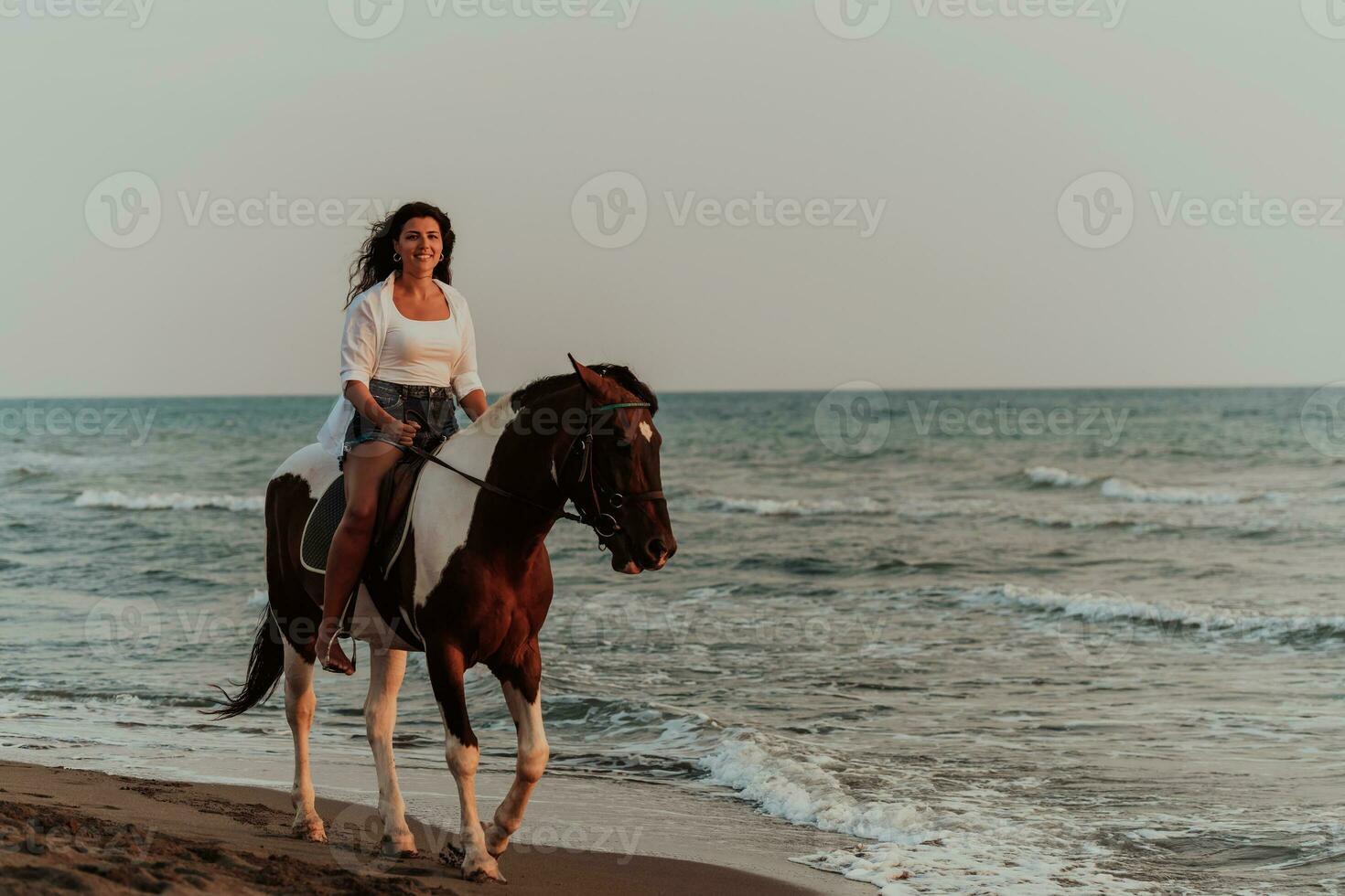 Woman in summer clothes enjoys riding a horse on a beautiful sandy beach at sunset. Selective focus photo