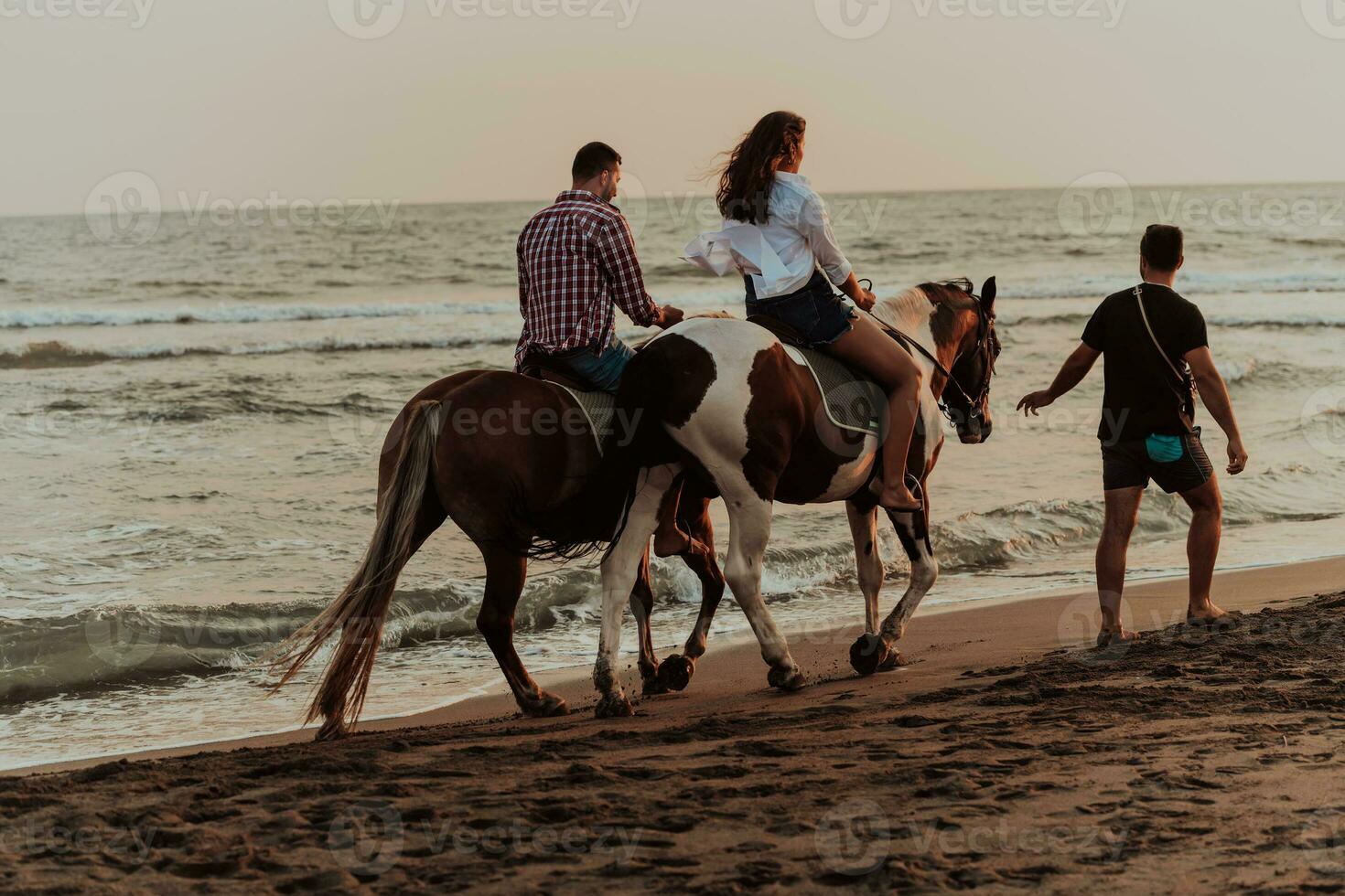 A loving couple in summer clothes riding a horse on a sandy beach at sunset. Sea and sunset in the background. Selective focus photo