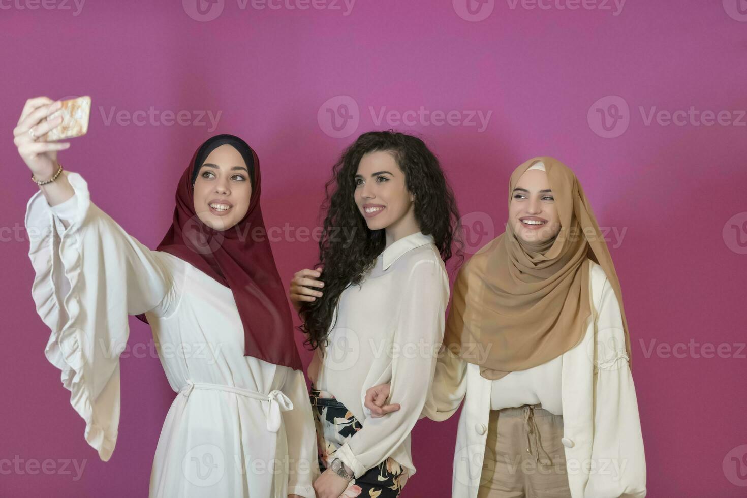 group of beautiful muslim women two of them in fashionable dress with hijab using mobile phone while taking selfie picture isolated on pink background representing modern islam fashion technology photo