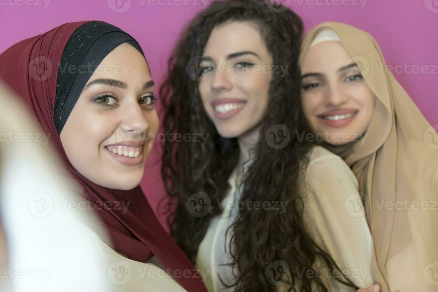 group of beautiful muslim women two of them in fashionable dress with hijab using mobile phone while taking selfie picture isolated on pink background representing modern islam fashion technology photo