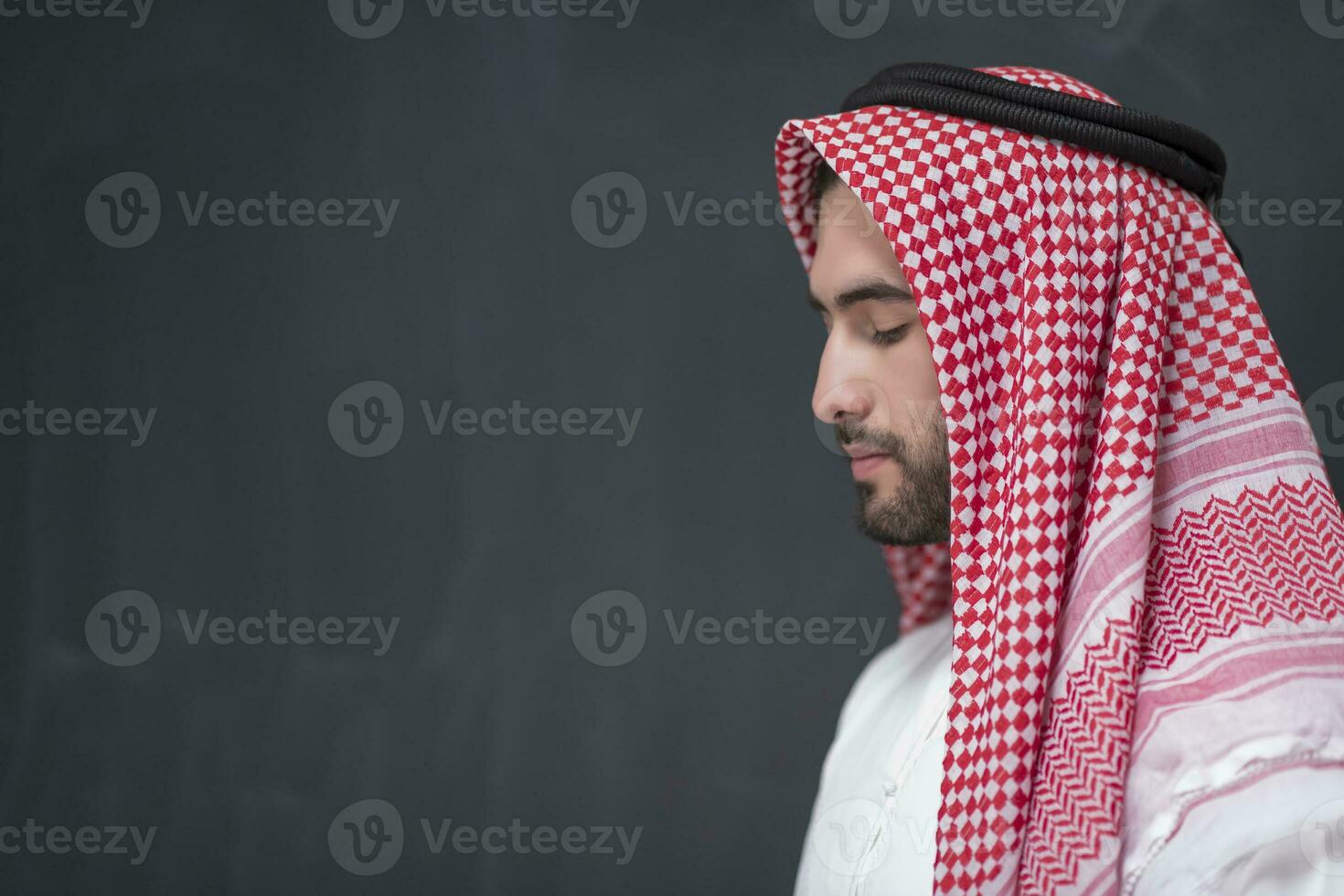 A young Arabian man in traditional clothes making a traditional prayer to God keeps his hands in praying gesture in front of a black background photo
