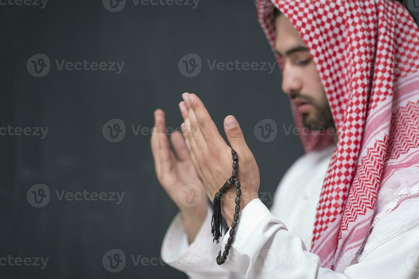 A young Arabian man in traditional clothes making a traditional prayer to God keeps his hands in praying gesture in front of a black background photo