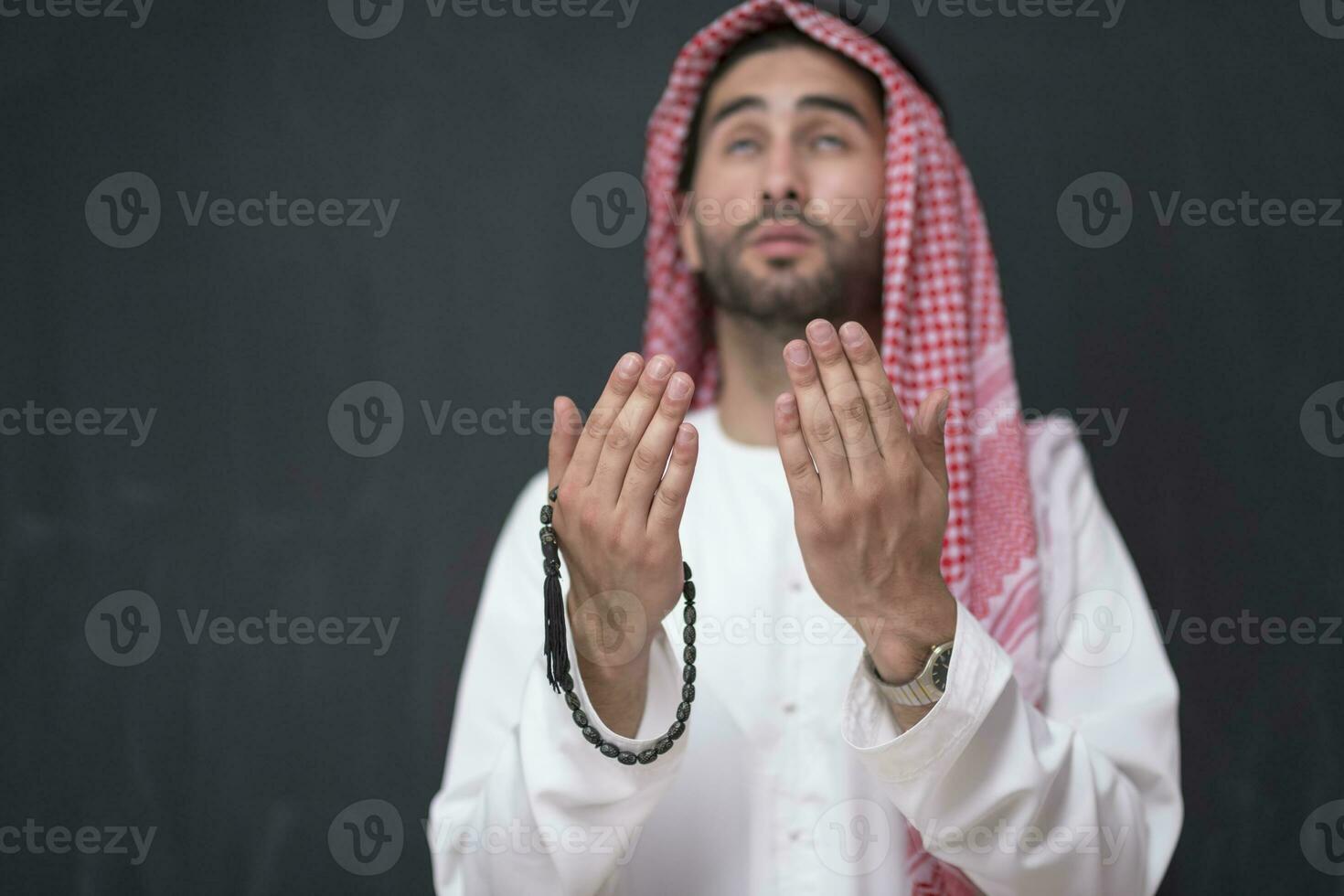 A young Arabian man in traditional clothes making a traditional prayer to God keeps his hands in praying gesture in front of a black background photo