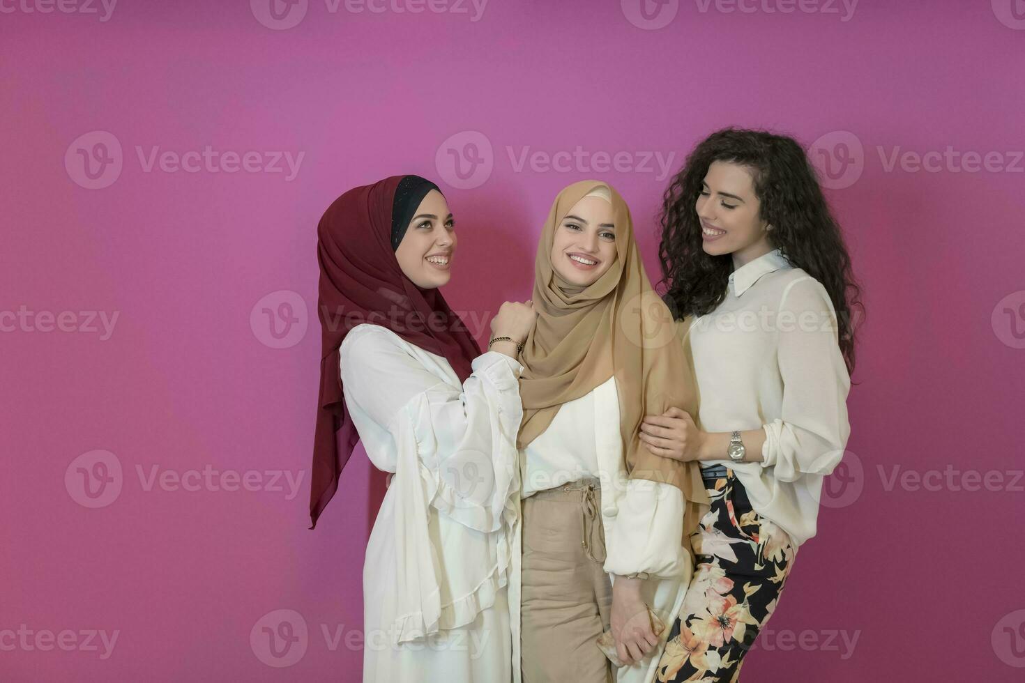 Group of beautiful muslim women two of them in fashionable dress with hijab using mobile phone isolated on pink background representing modern islam fashion technology and ramadan kareem concept photo