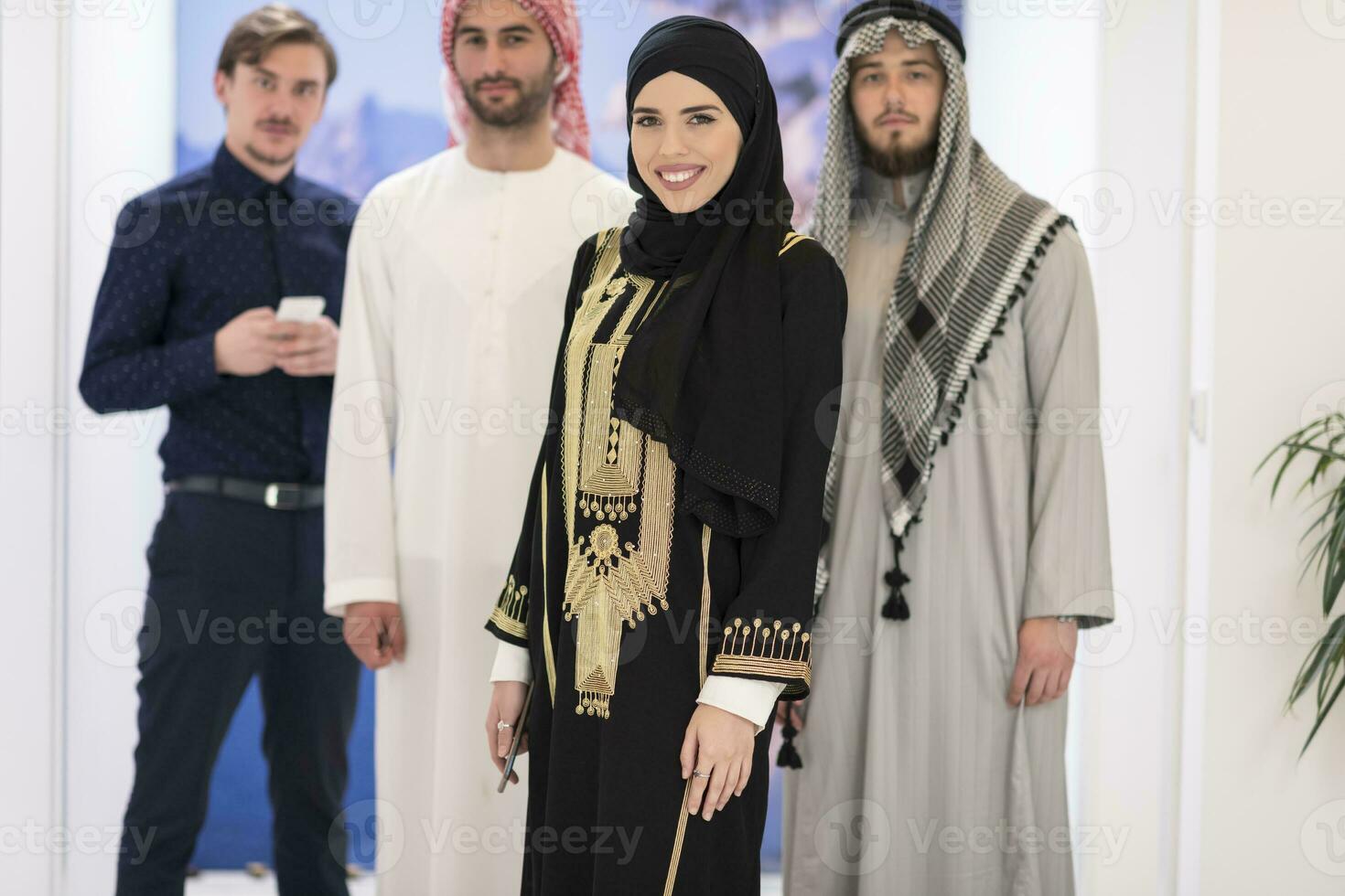 Group portrait of young Muslim people women in fashionable hijab dress with three Arabian men using smartphones at modern bright home photo