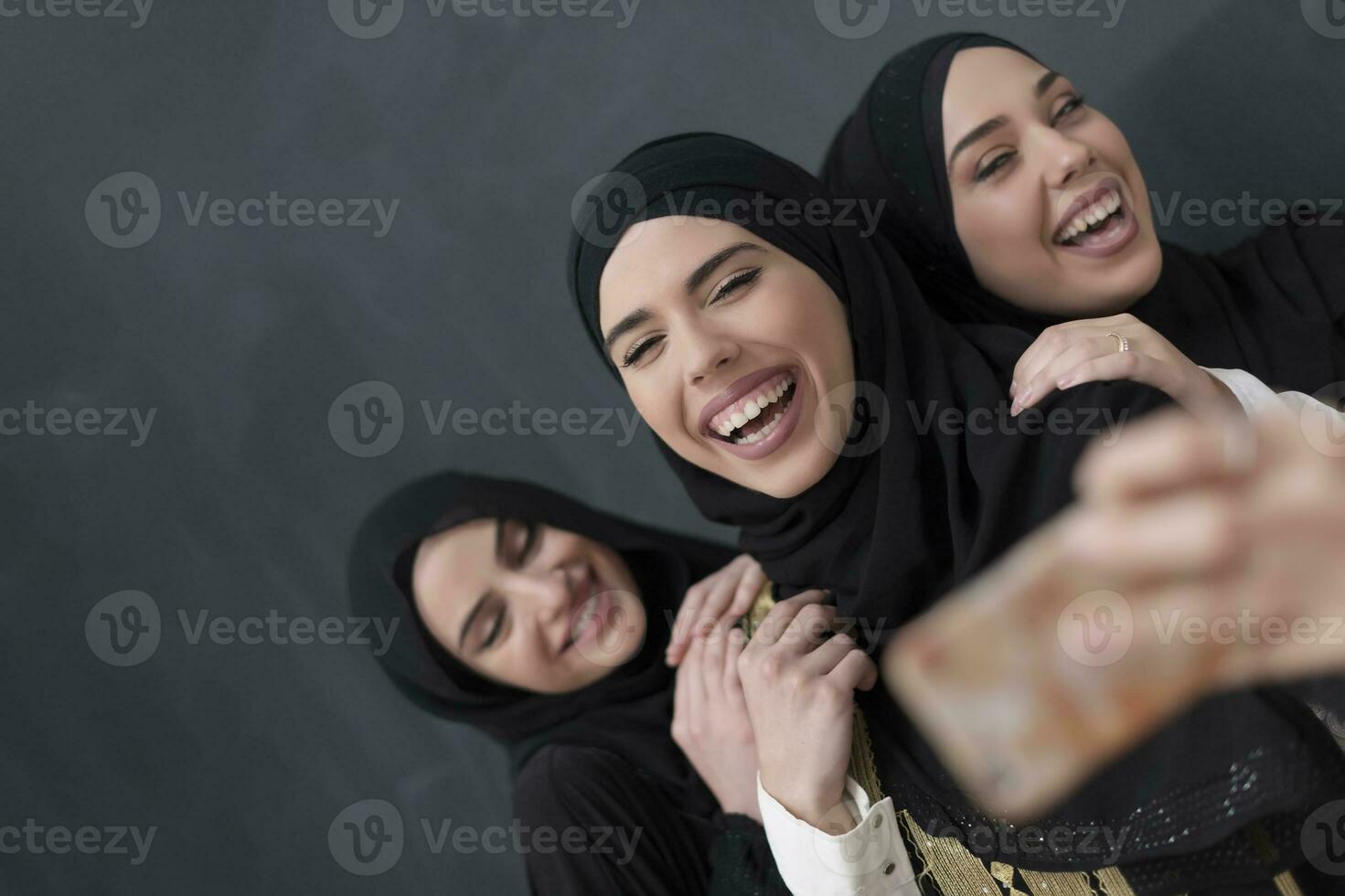 Group of young beautiful muslim women in fashionable dress with hijab using smartphone while taking selfie picture in front of black background photo