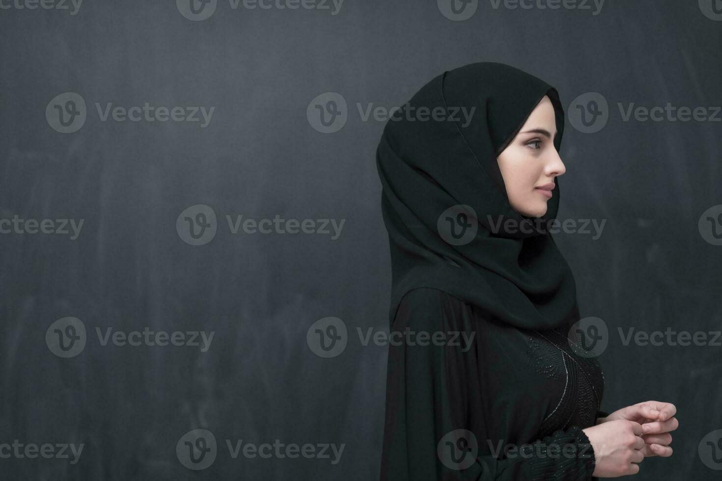 Portrait of beautiful muslim woman in fashionable dress with hijab in front of black chalkboard representing modern islam fashion and ramadan kareem concept photo