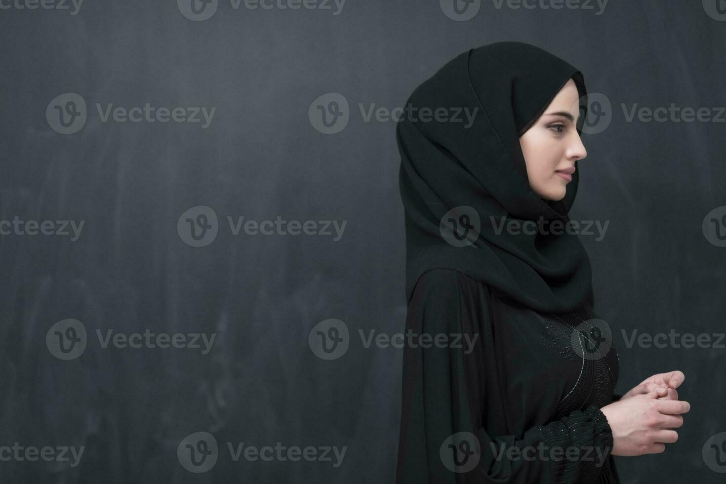 Portrait of beautiful muslim woman in fashionable dress with hijab in front of black chalkboard representing modern islam fashion and ramadan kareem concept photo