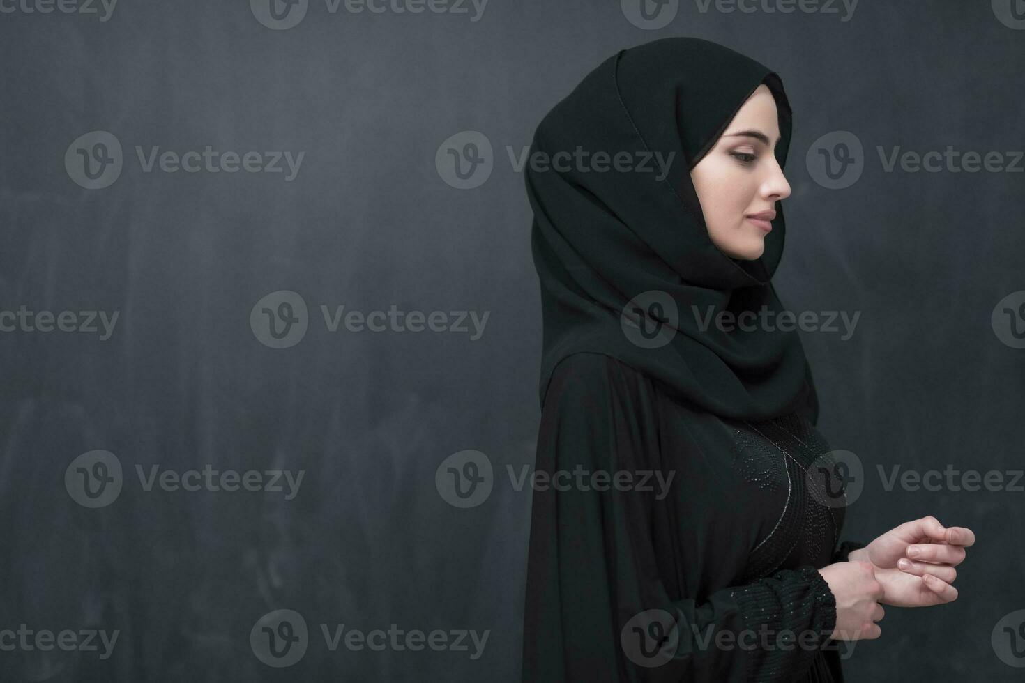 Portrait of beautiful muslim woman in fashionable dress with hijab in front of black chalkboard representing modern islam fashion and ramadan kareem concept photo