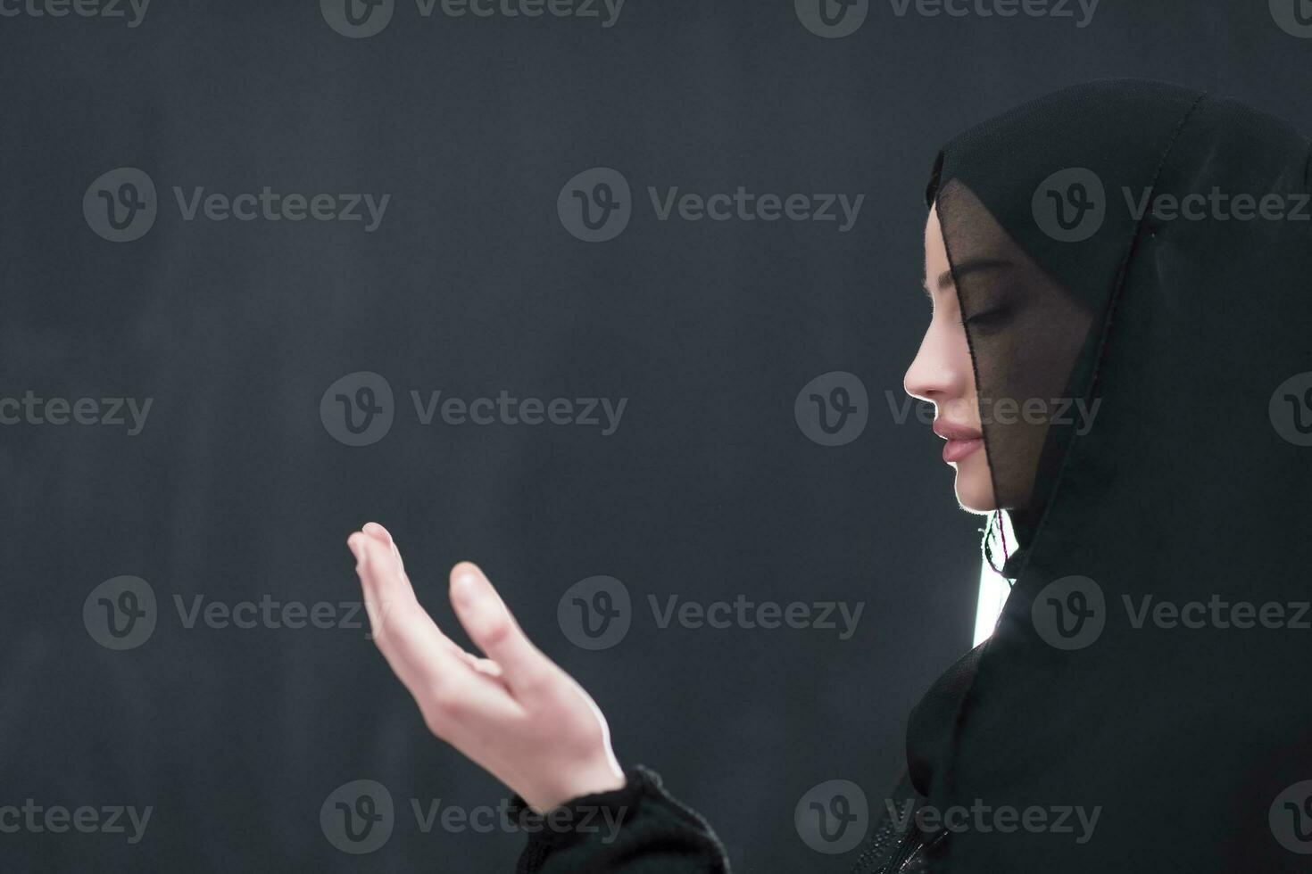 Portrait of beautiful muslim woman in fashionable dress with hijab making traditional prayer to God keeps hands in praying gesture isolated on black background photo