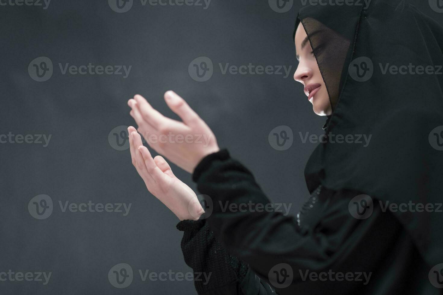 Portrait of beautiful muslim woman in fashionable dress with hijab making traditional prayer to God keeps hands in praying gesture isolated on black background photo