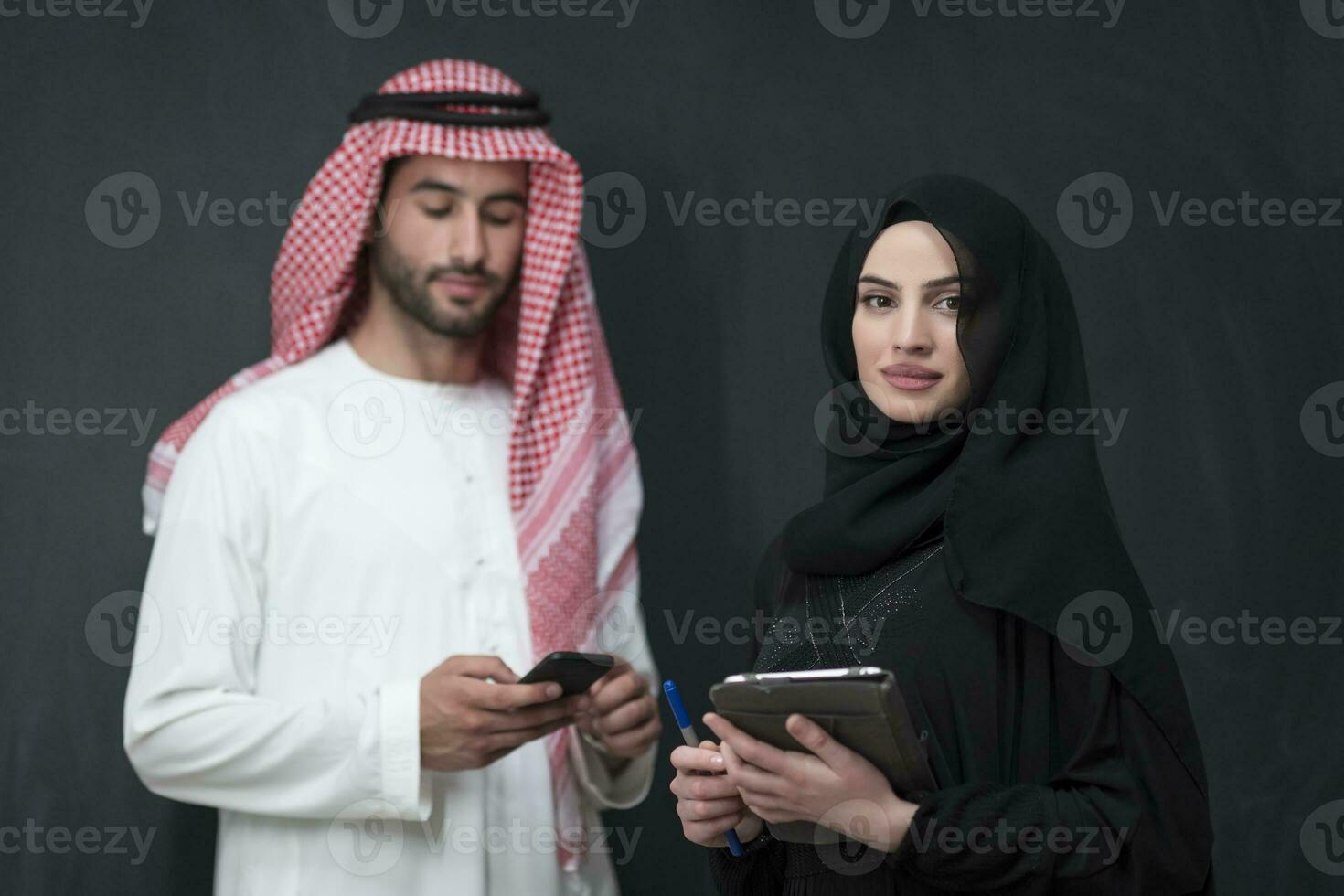 Young muslim business couple in fashionable hijab dress using smartphone and tablet in front of black background photo