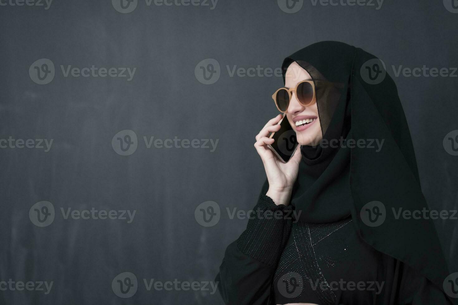Young modern muslim businesswoman using smartphone wearing sunglasses and hijab clothes in front of black chalkboard photo