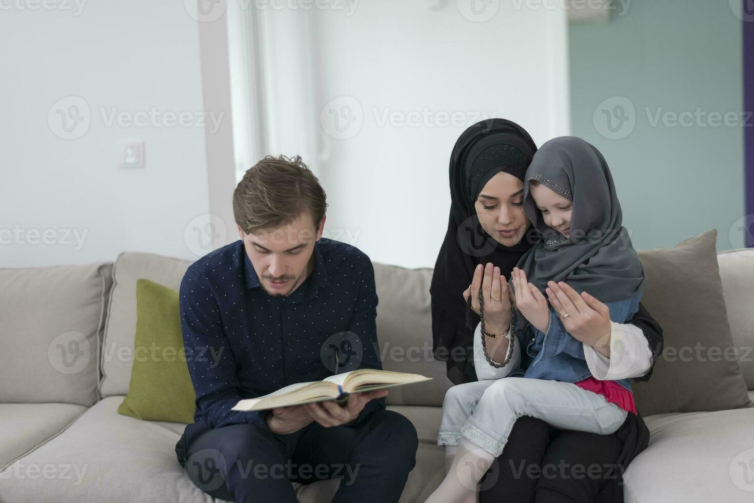 tradicional musulmán familia padres con niños leyendo Corán y Orando juntos en el sofá antes de iftar cena durante un Ramadán banquete a hogar foto