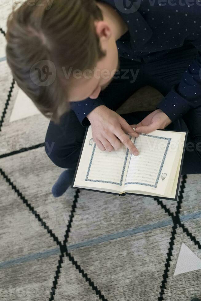 joven musulmán hombre sentado en el piso mientras leyendo santo libro Corán antes de iftar cena durante un Ramadán banquete a hogar foto