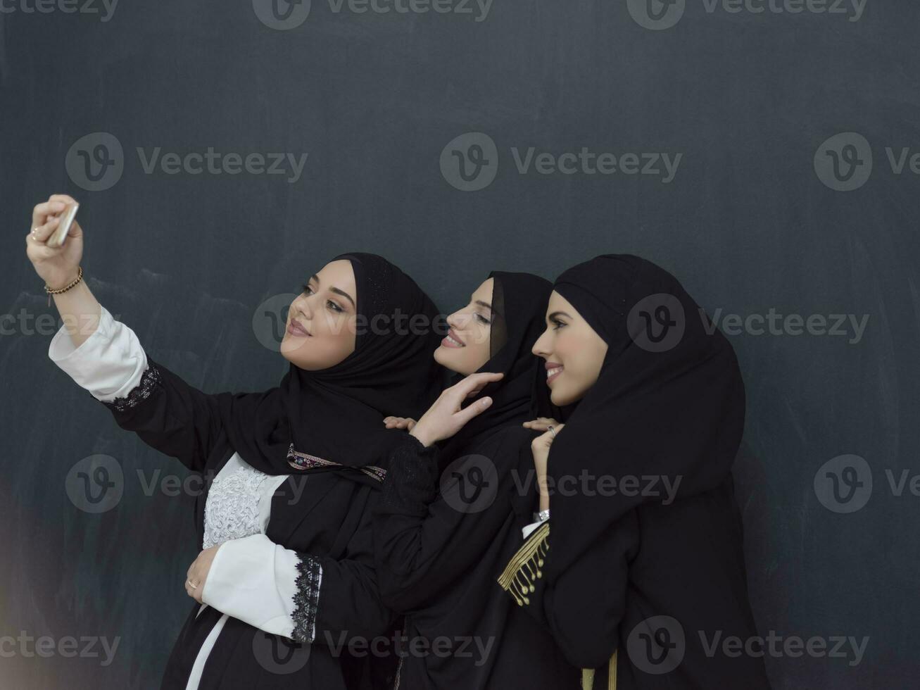Group of young muslim women in fashionable dress with hijab using smartphone while taking selfie picture in front of black background photo