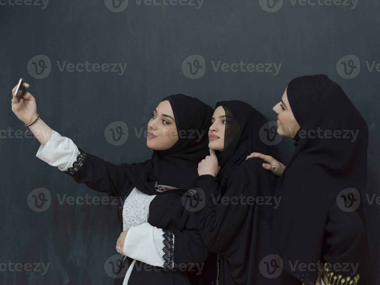 Group of young muslim women in fashionable dress with hijab using smartphone while taking selfie picture in front of black background photo