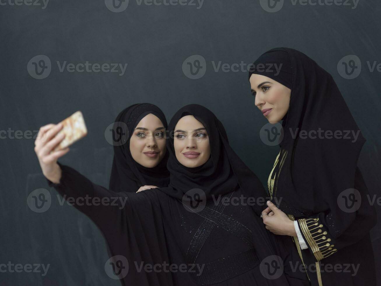 Group of young muslim women in fashionable dress with hijab using smartphone while taking selfie picture in front of black background photo