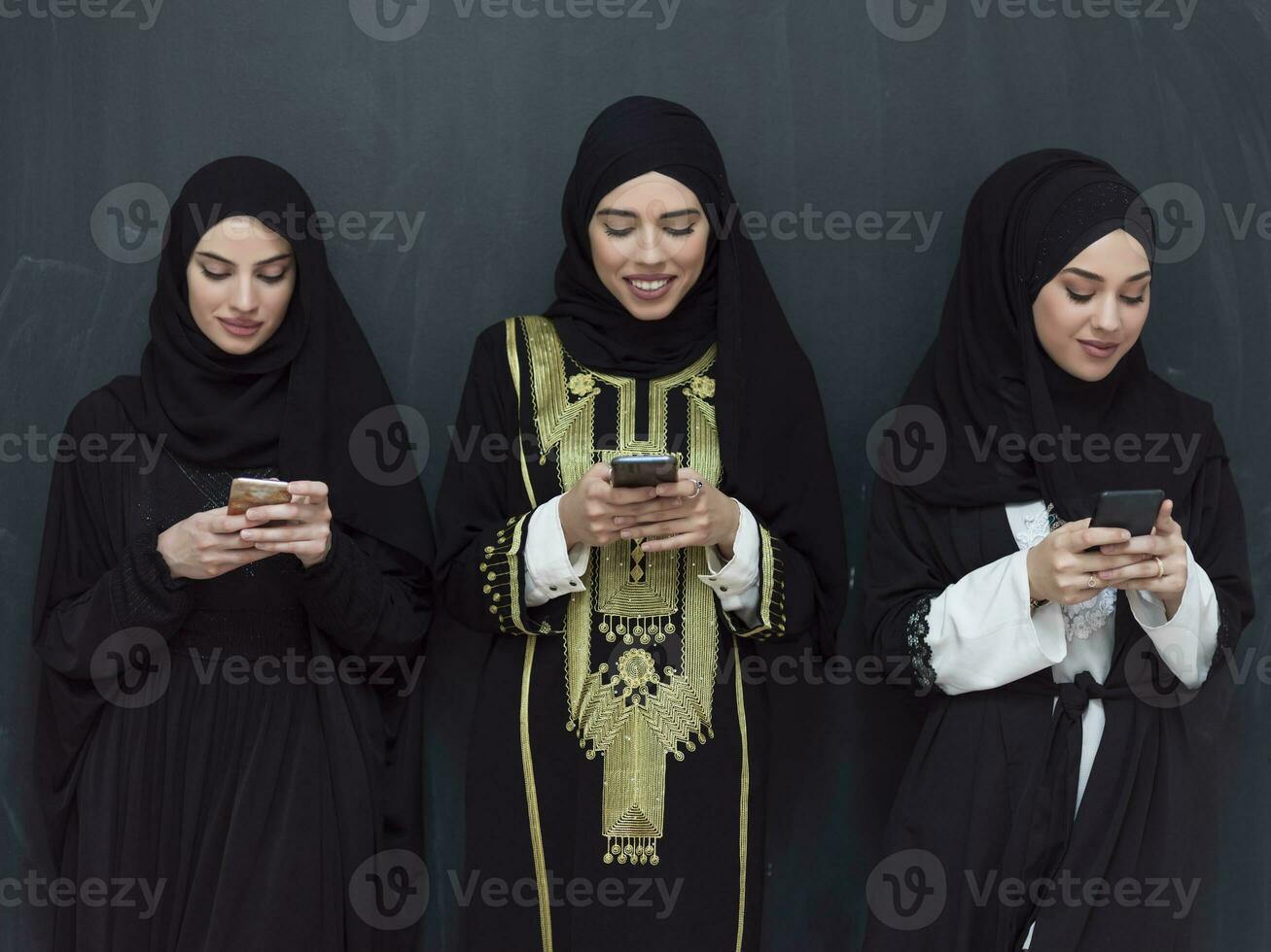 group of beautiful muslim women in fashionable dress with hijab using mobile phone in front of black chalkboard representing modern islam fashion technology and ramadan kareem concept photo