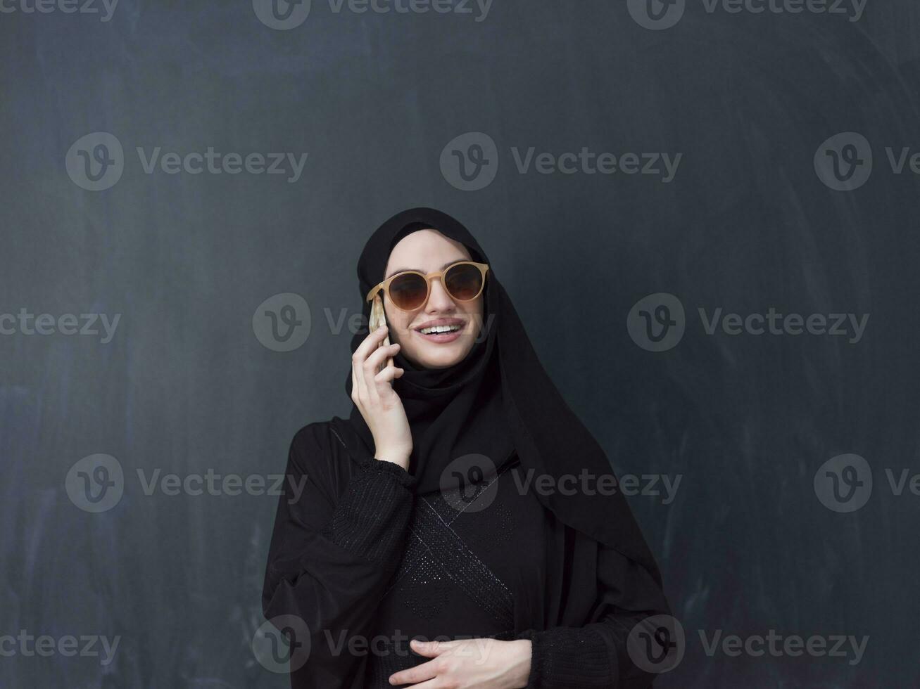 young modern muslim business woman using smartphone wearing sunglasses and hijab clothes in front of black chalkboard photo