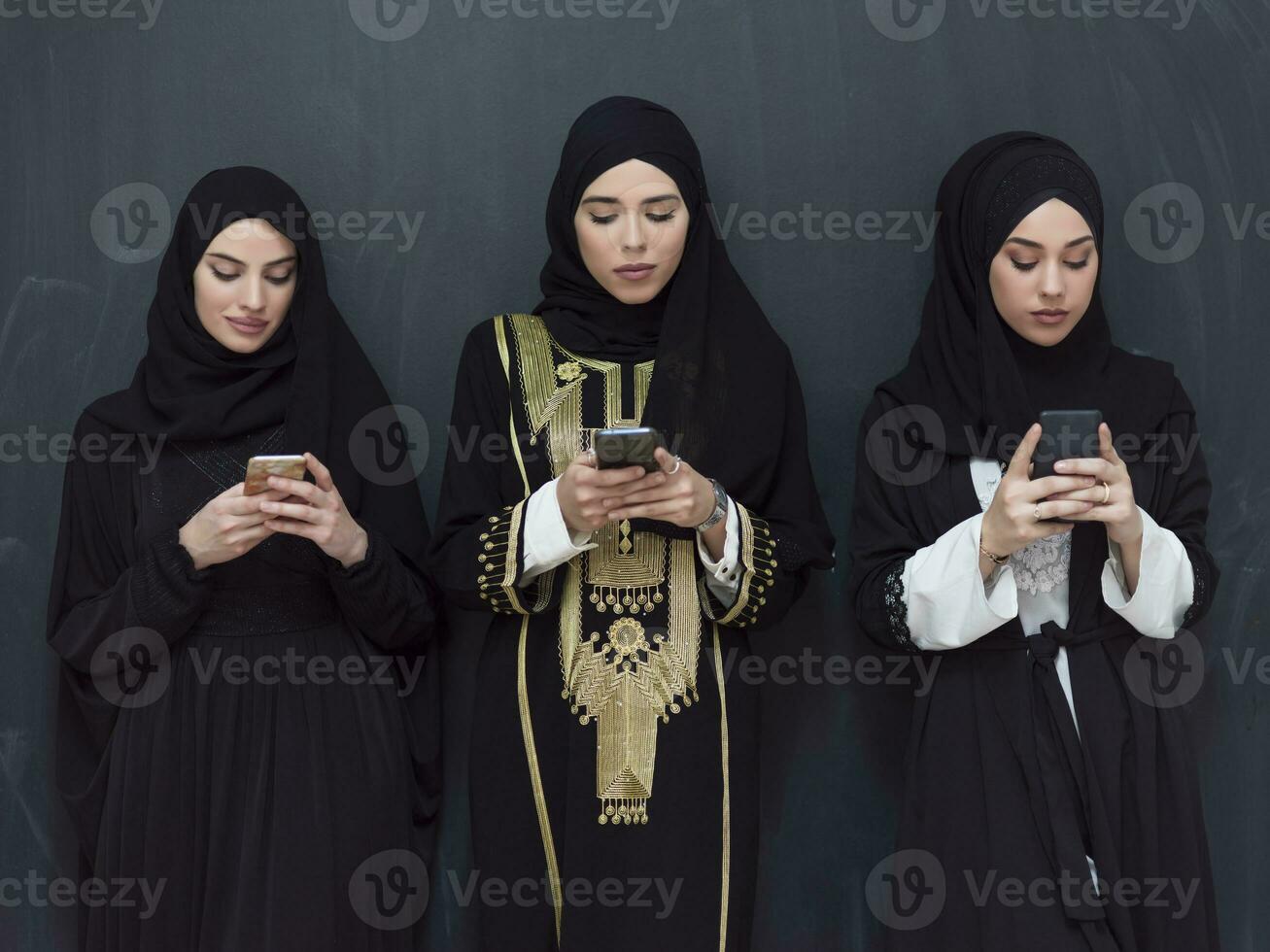 group of beautiful muslim women in fashionable dress with hijab using mobile phone in front of black chalkboard representing modern islam fashion technology and ramadan kareem concept photo