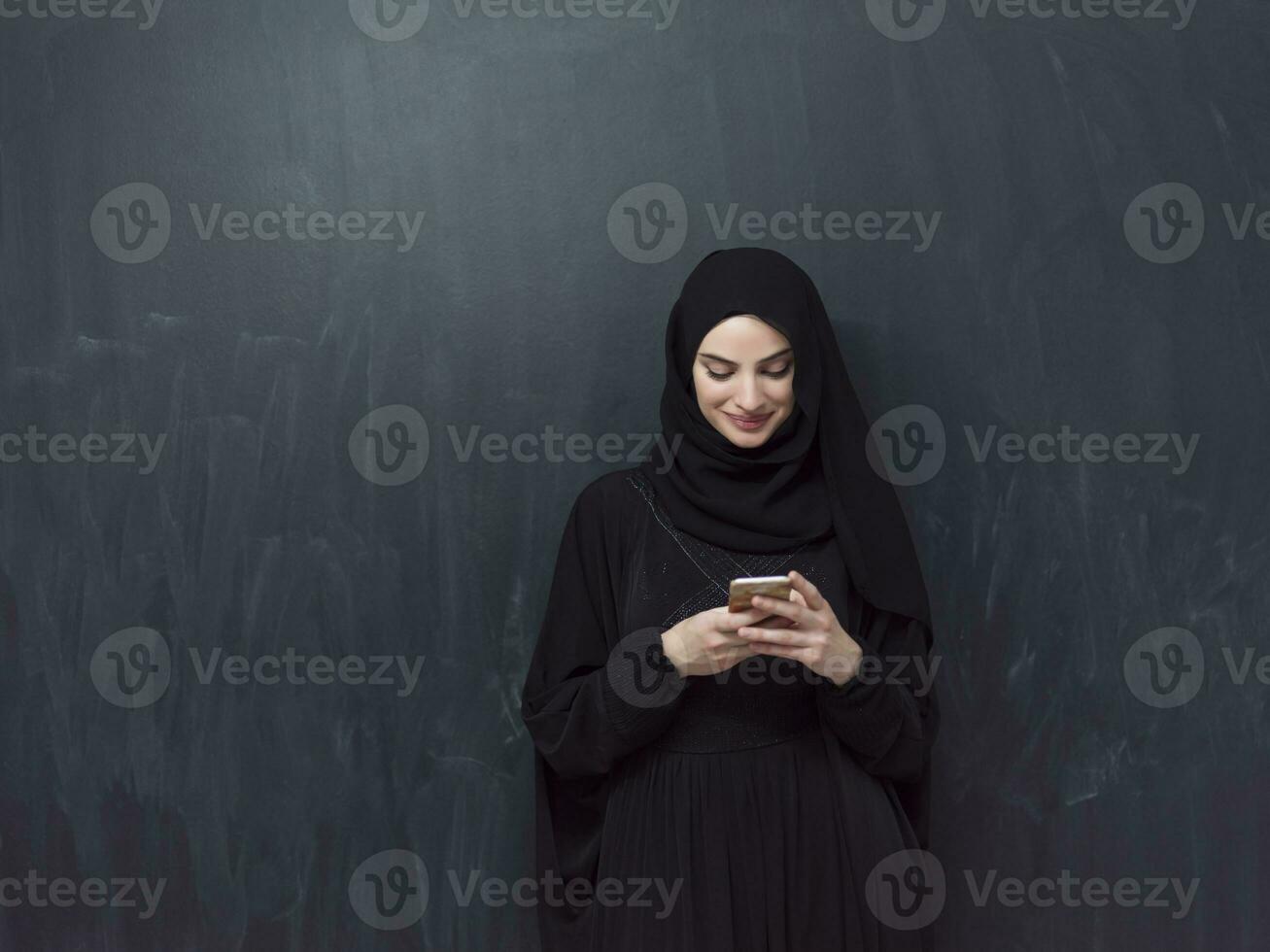 Young modern muslim business woman using smartphone wearing hijab clothes in front of black chalkboard photo