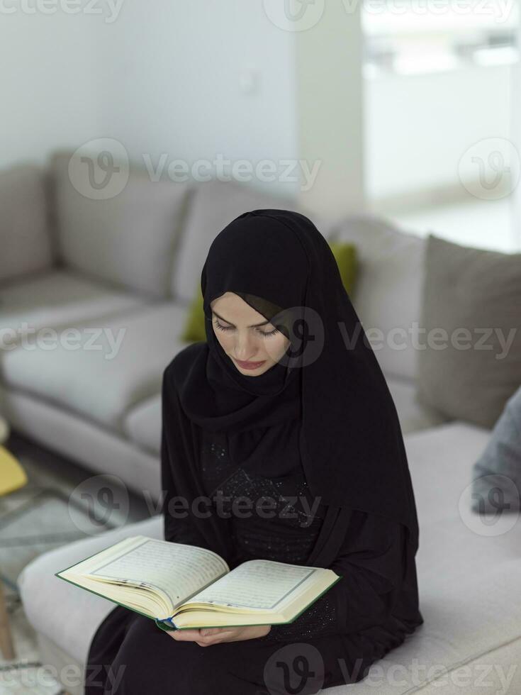 Young traditional muslim woman reading Quran on the sofa before iftar dinner during a ramadan feast at home photo