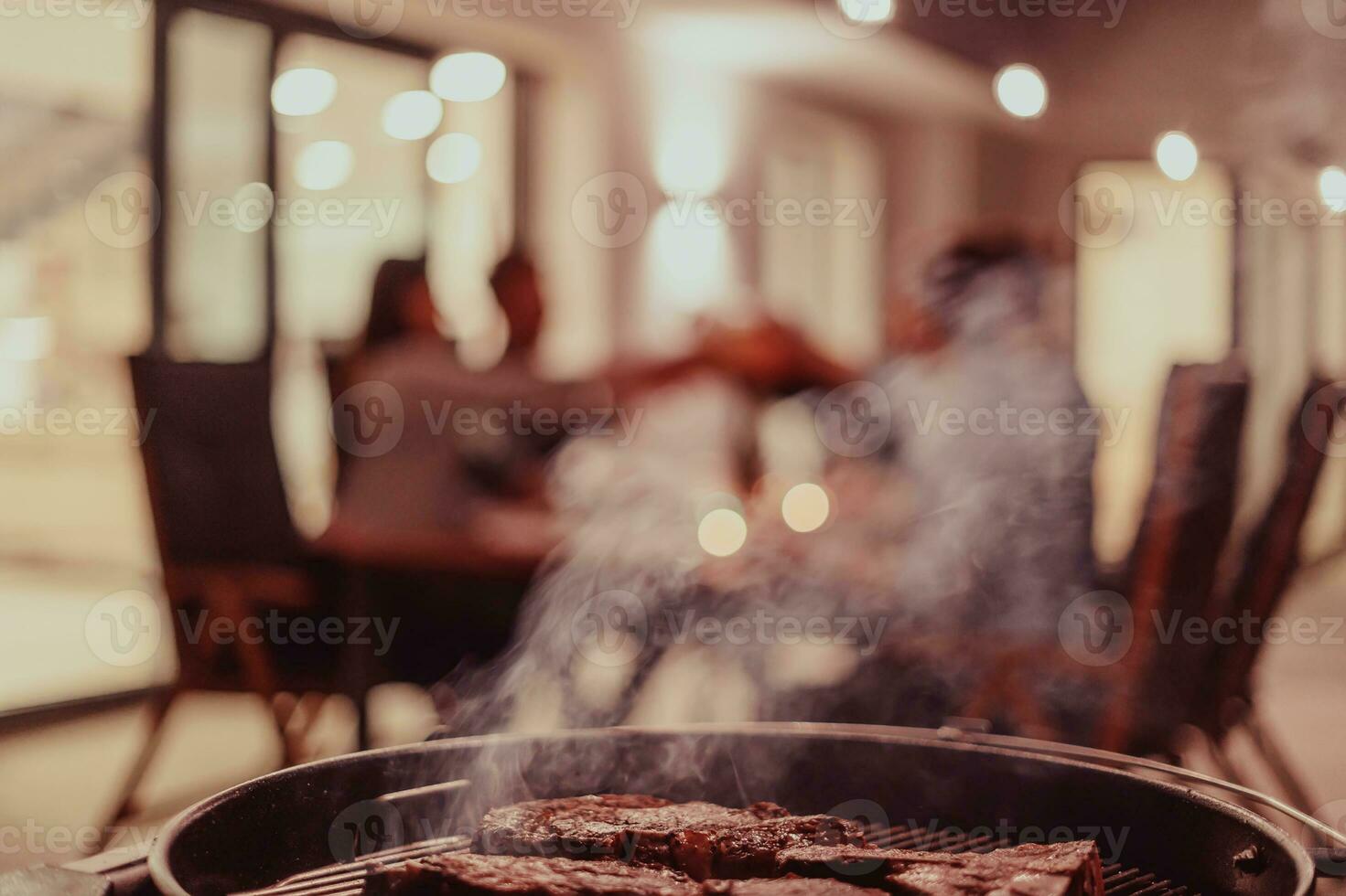 Close-up photo of delicious meat being grilled. In the background, friends and family are sitting and waiting for a meal