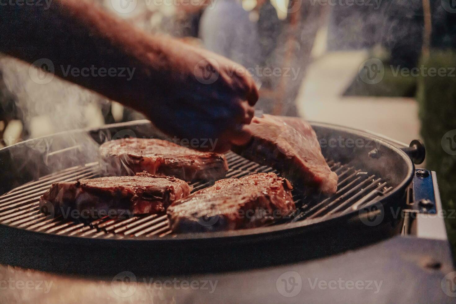 Close-up photo of delicious meat being grilled. In the background, friends and family are sitting and waiting for a meal