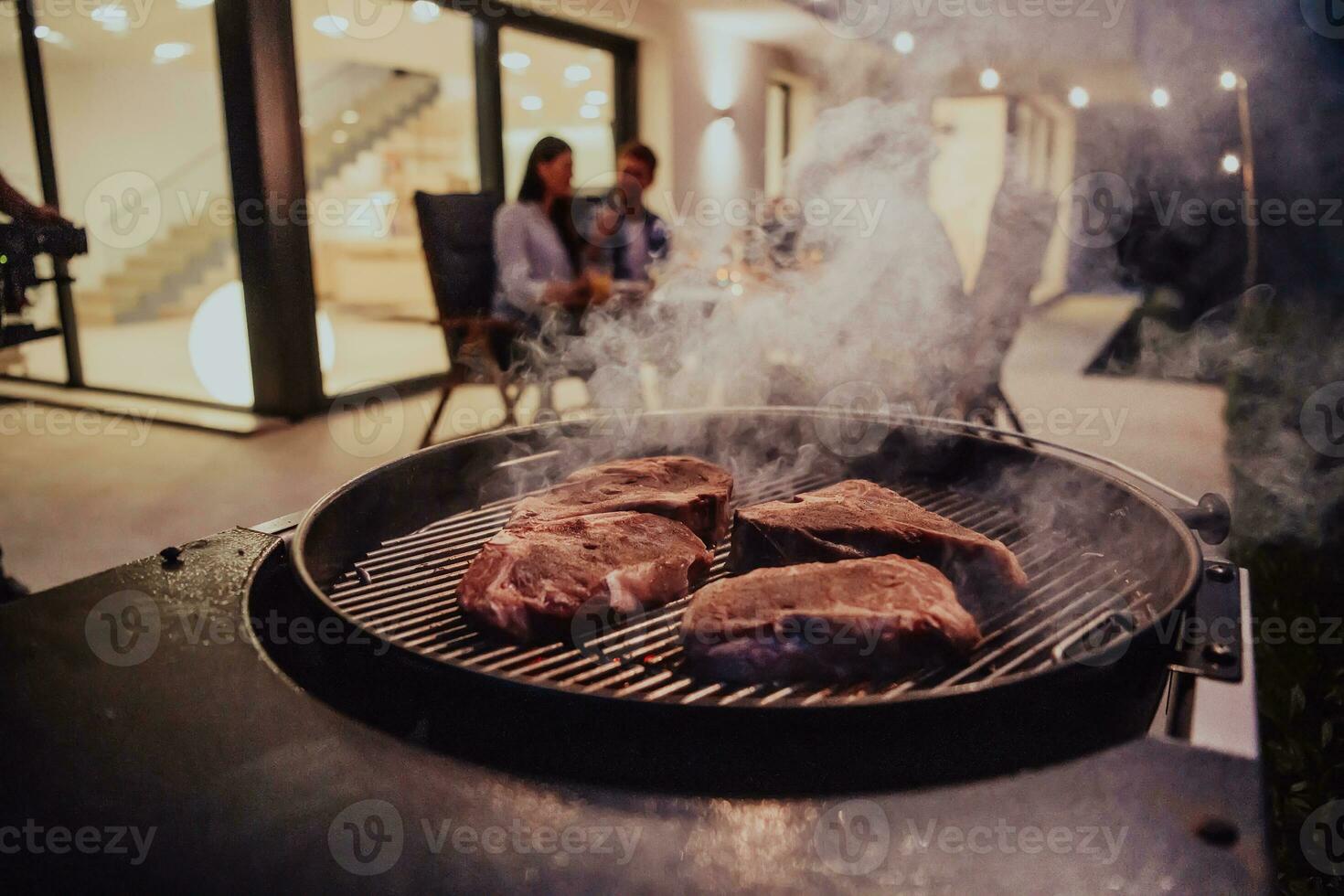 Close-up photo of delicious meat being grilled. In the background, friends and family are sitting and waiting for a meal