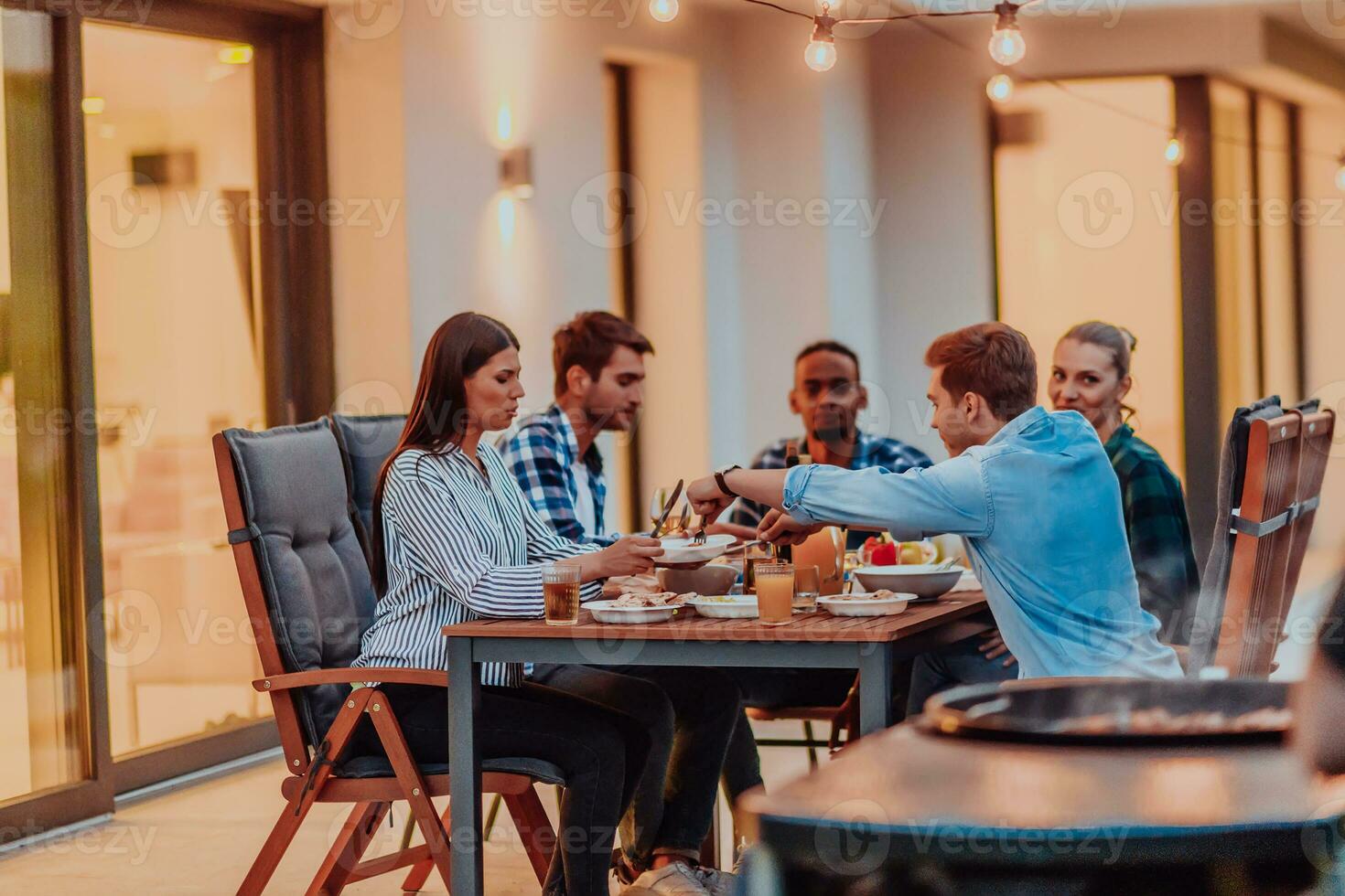 A group of young diverse people having dinner on the terrace of a modern house in the evening. Fun for friends and family. Celebration of holidays, weddings with barbecue. photo