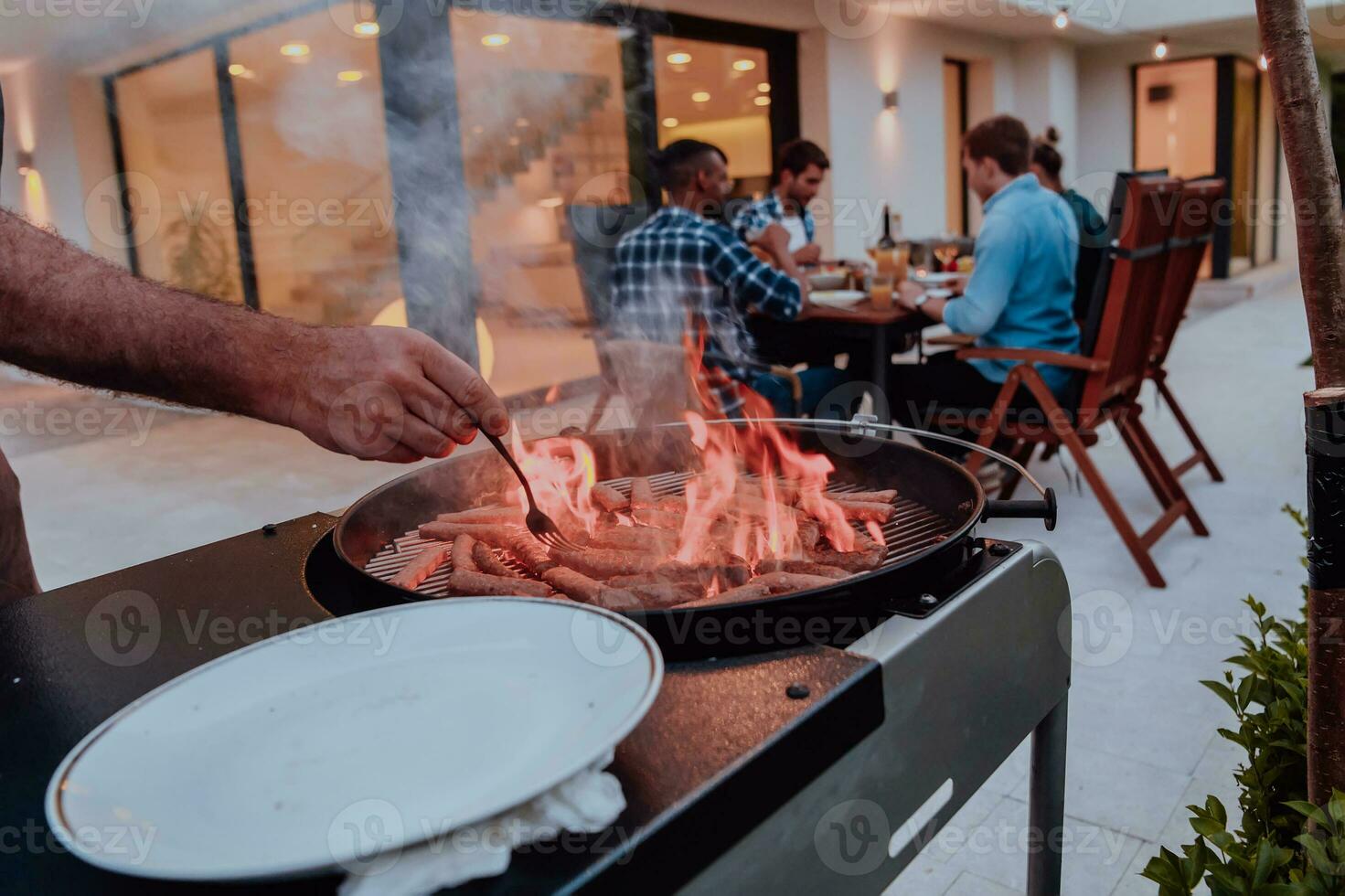 A group of friends and family barbecue together in the evening on the terrace in front of a large modern house photo