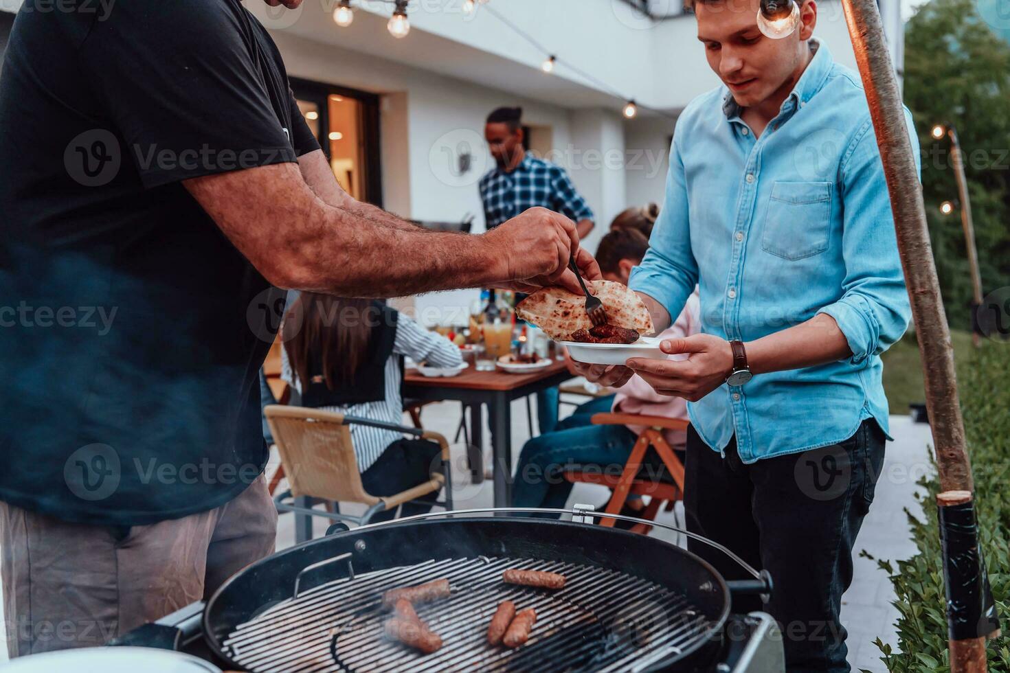 A group of friends and family barbecue together in the evening on the terrace in front of a large modern house photo