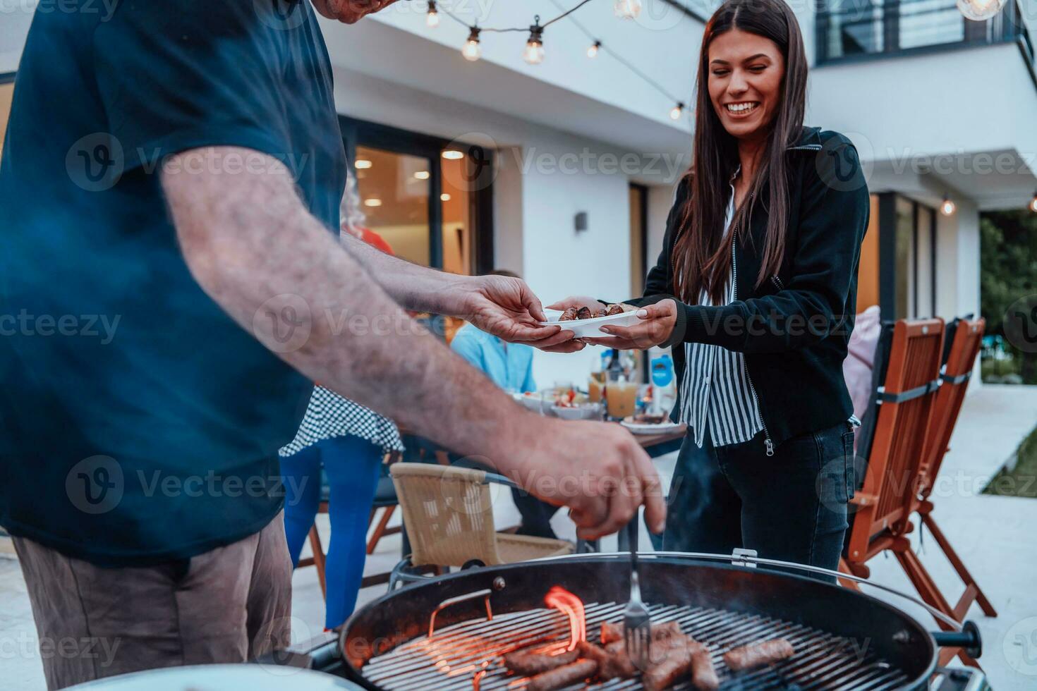 A group of friends and family barbecue together in the evening on the terrace in front of a large modern house photo