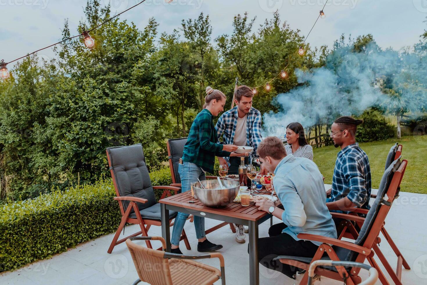 un grupo de joven diverso personas teniendo cena en el terraza de un moderno casa en el noche. divertido para amigos y familia. celebracion de vacaciones, bodas con parilla. foto