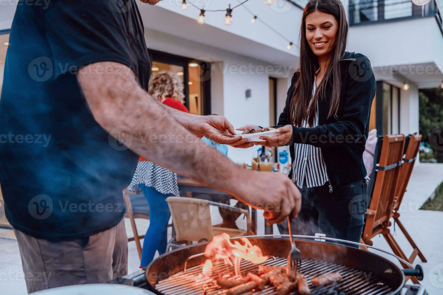 A group of friends and family barbecue together in the evening on the terrace in front of a large modern house photo