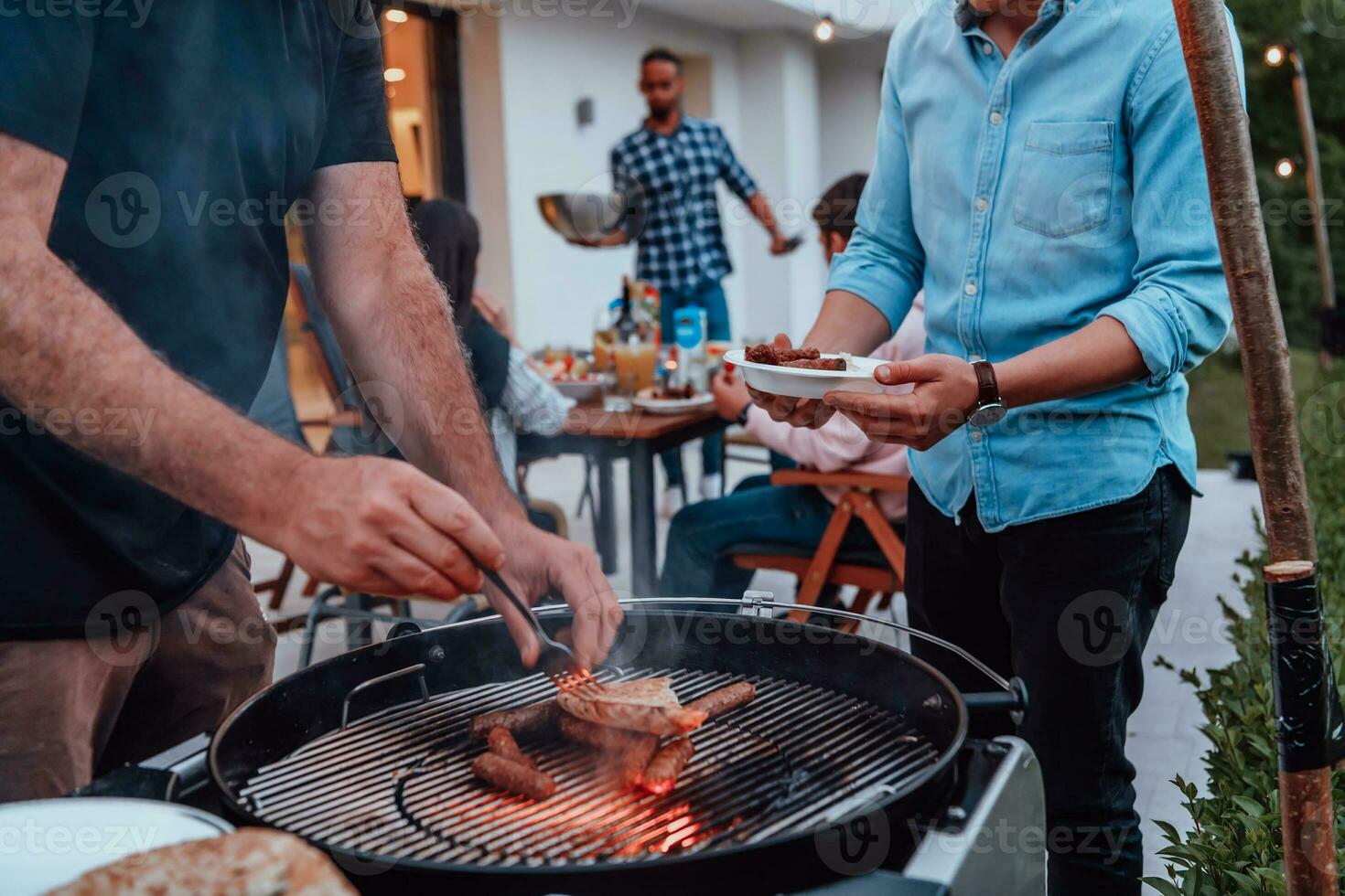 un grupo de amigos y familia parilla juntos en el noche en el terraza en frente de un grande moderno casa foto