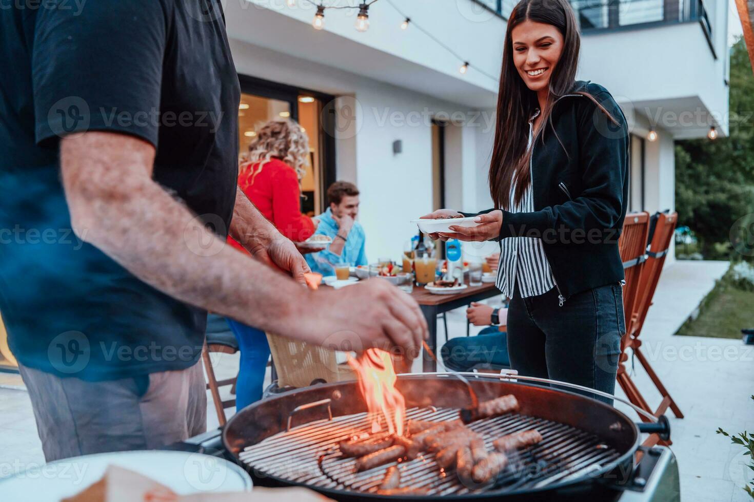 A group of friends and family barbecue together in the evening on the terrace in front of a large modern house photo